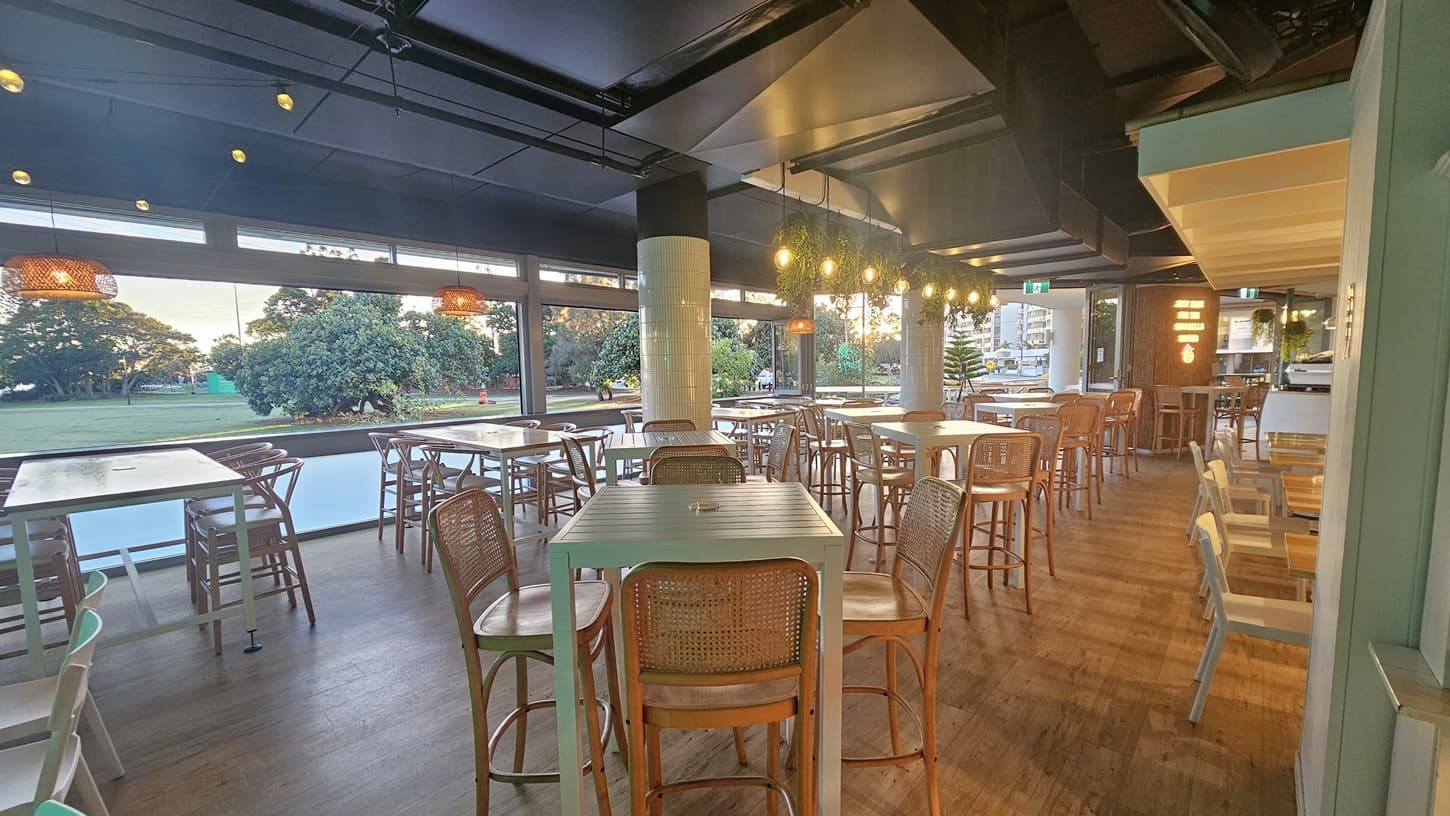 On the left, there are wooden tables with matching chairs, positioned near large open windows. In the middle, the background features a grassy park with trees and people, and a road with parked cars. On the right, more wooden tables and chairs are near another large open window. The floor is wooden, and the ceiling has dark panels with two hanging wicker lights.