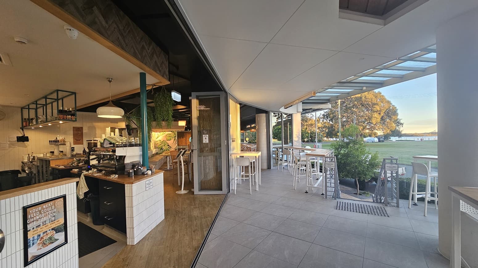 On the left, there is an outdoor seating area with wooden tables and chairs under a covered patio. The right side features an open café with a counter, espresso machine, and shelves stocked with bottles and jars. In the middle, there is a large glass door that can be folded open, connecting the indoor and outdoor spaces. The background includes more seating and greenery. The floor is a mix of light wood and large grey tiles, and the ceiling is white with recessed lighting and wooden panels.
