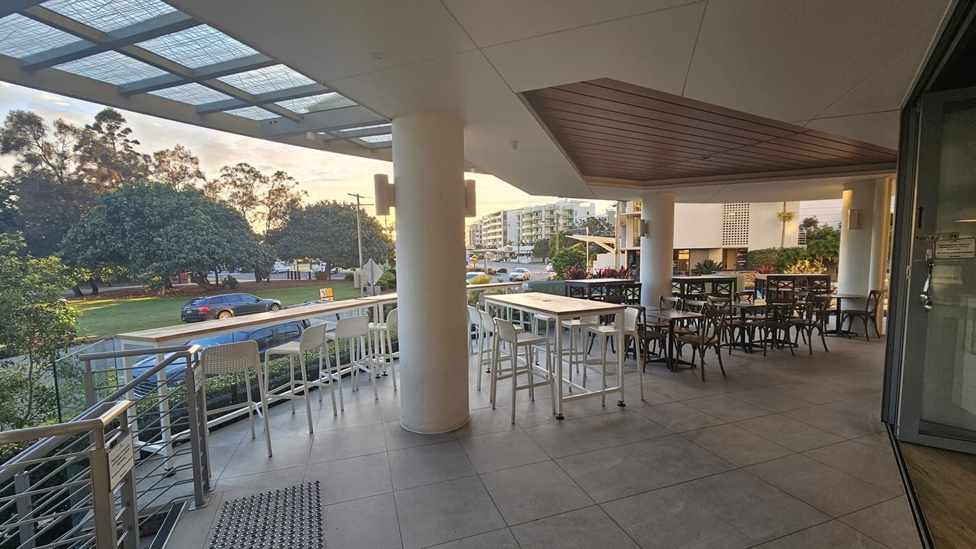 On the left, there are tall white bar stools lined up against a railing, with a view of trees and a road in the background. In the middle, a large white column supports the ceiling, and there are more tables and chairs. On the right, there are additional tables and chairs, with a glass door leading inside. The floor is tiled in grey, and the ceiling has a wooden panel section above the seating area.