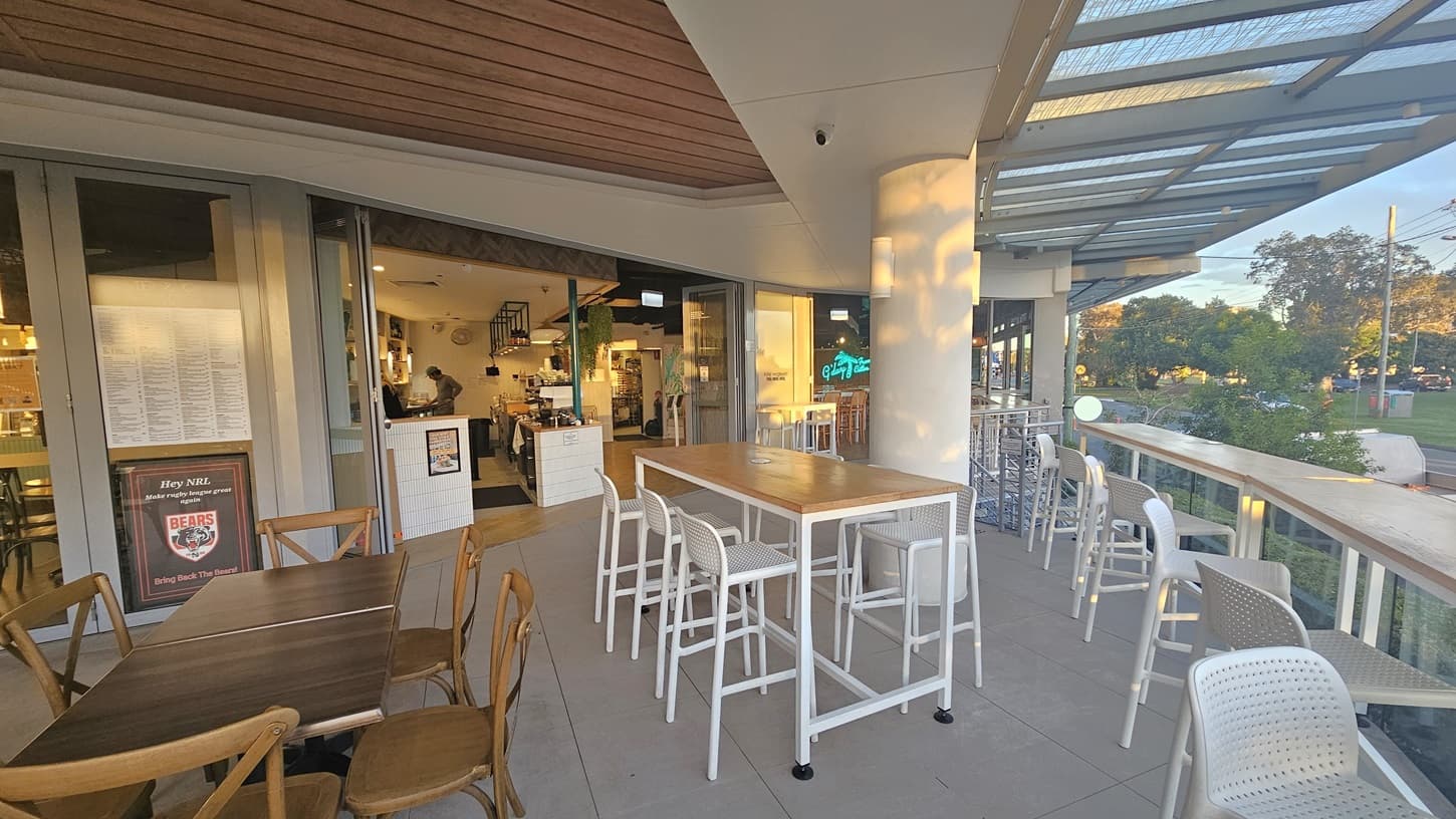 On the left, there are wooden tables with matching chairs, and a menu board is visible near the entrance. In the middle, a tall white column stands next to a high table with white bar stools. On the right, there is a glass railing with a view of trees and a road. The floor is tiled in light grey, and the ceiling has wooden panels above the seating area and a glass canopy extending outward.