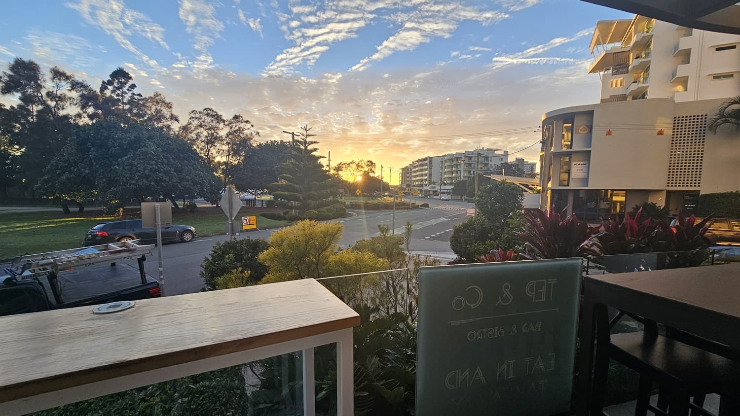 On the left, there is a wooden table with a white frame, positioned near a glass railing. In the middle, the background features a road with cars, trees, and buildings, with the sun setting behind them. On the right, there is a glass panel with text, and beyond it, more buildings and greenery.