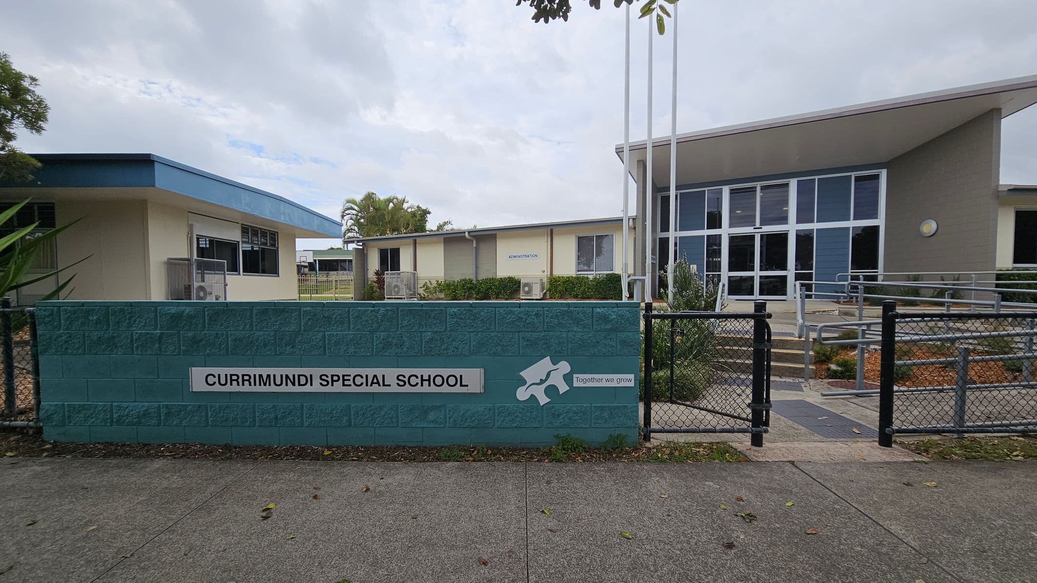 A school entrance with a blue brick wall in the foreground displaying the sign "Currimundi Special School" and the phrase "Together we grow" alongside a puzzle piece graphic. Behind the wall, there are two buildings. The left building has a light green exterior with a blue roof, while the right building features large glass windows and a sloped roof. A pathway with railings leads to the entrance. The ground is paved, and there are some plants and trees visible.