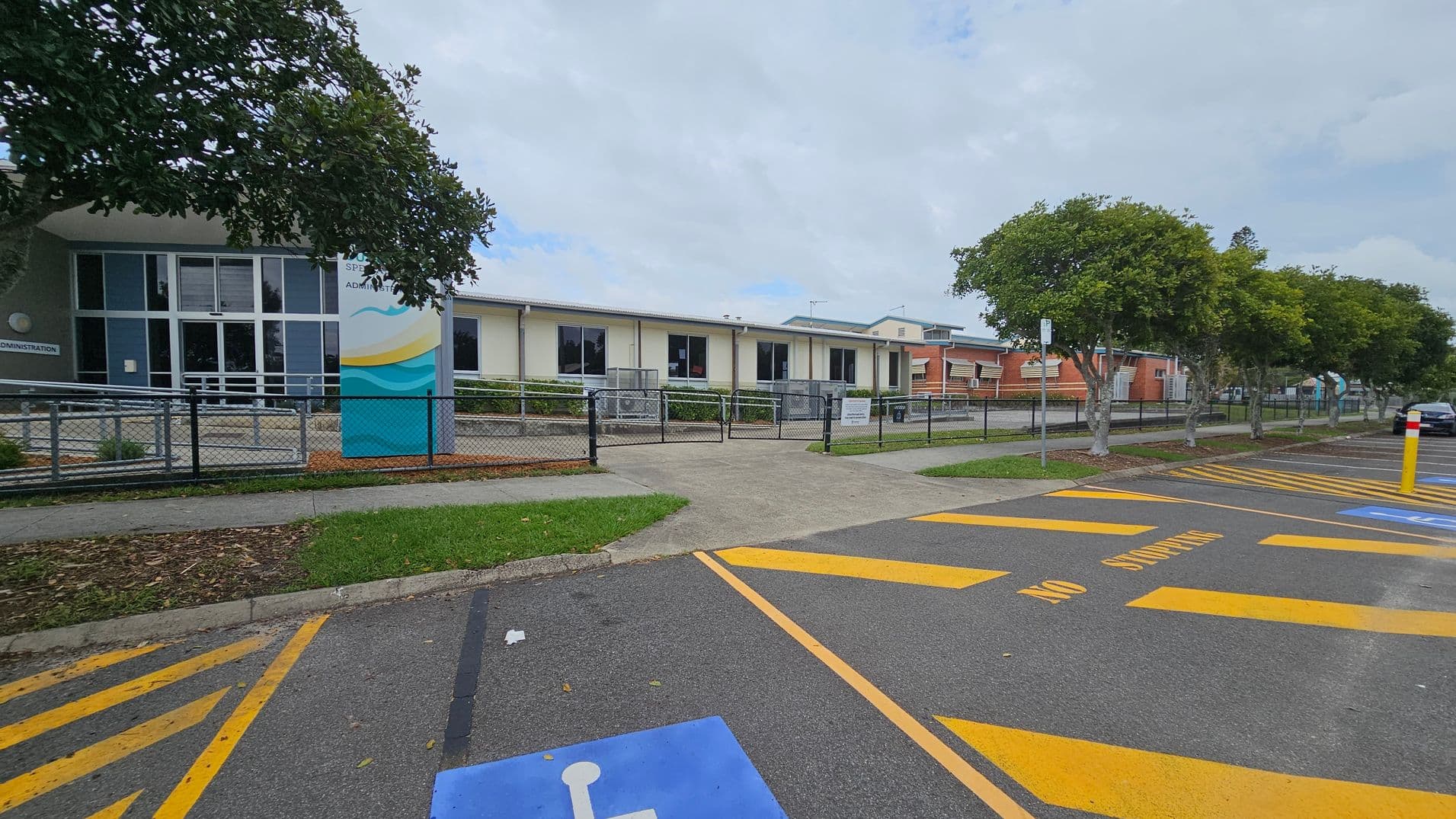 A school building with a modern design is in the background, featuring large windows and a sign near the entrance. A fence runs along the front, with a pathway leading to the entrance. Trees line the right side, providing some greenery. In the foreground, a car park is visible with marked spaces, including a blue disabled parking spot and yellow "No Stopping" lines. The ground is asphalt, and the area is well-maintained.