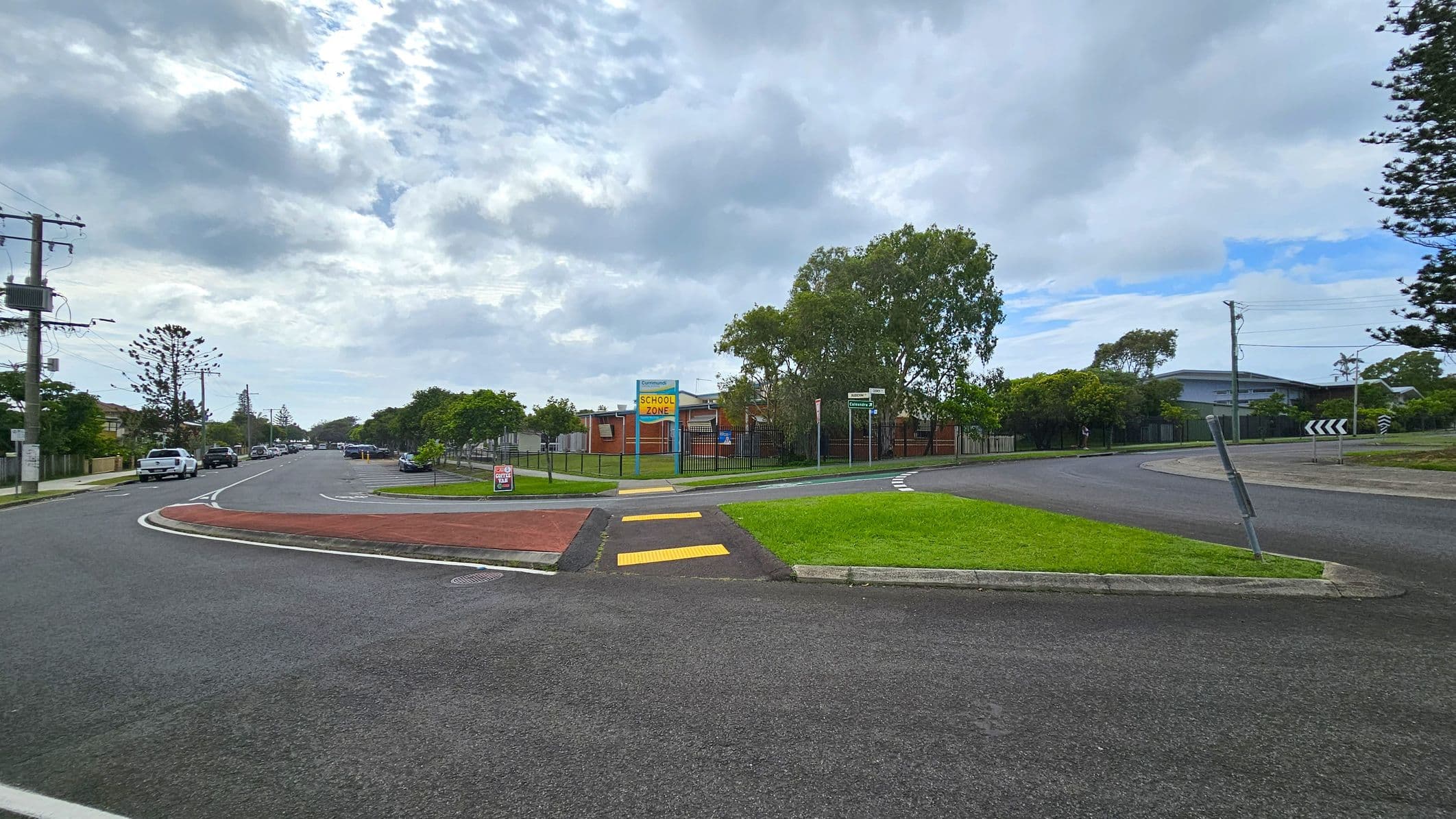 A street scene with a roundabout in the foreground. On the left, a road lined with parked cars and utility poles extends into the distance. In the centre, a grassy area with a "School Zone" sign and a pedestrian crossing with yellow markings. To the right, a curved road with a roundabout sign and trees in the background. A building is partially visible behind the trees. The sky is overcast with patches of blue.