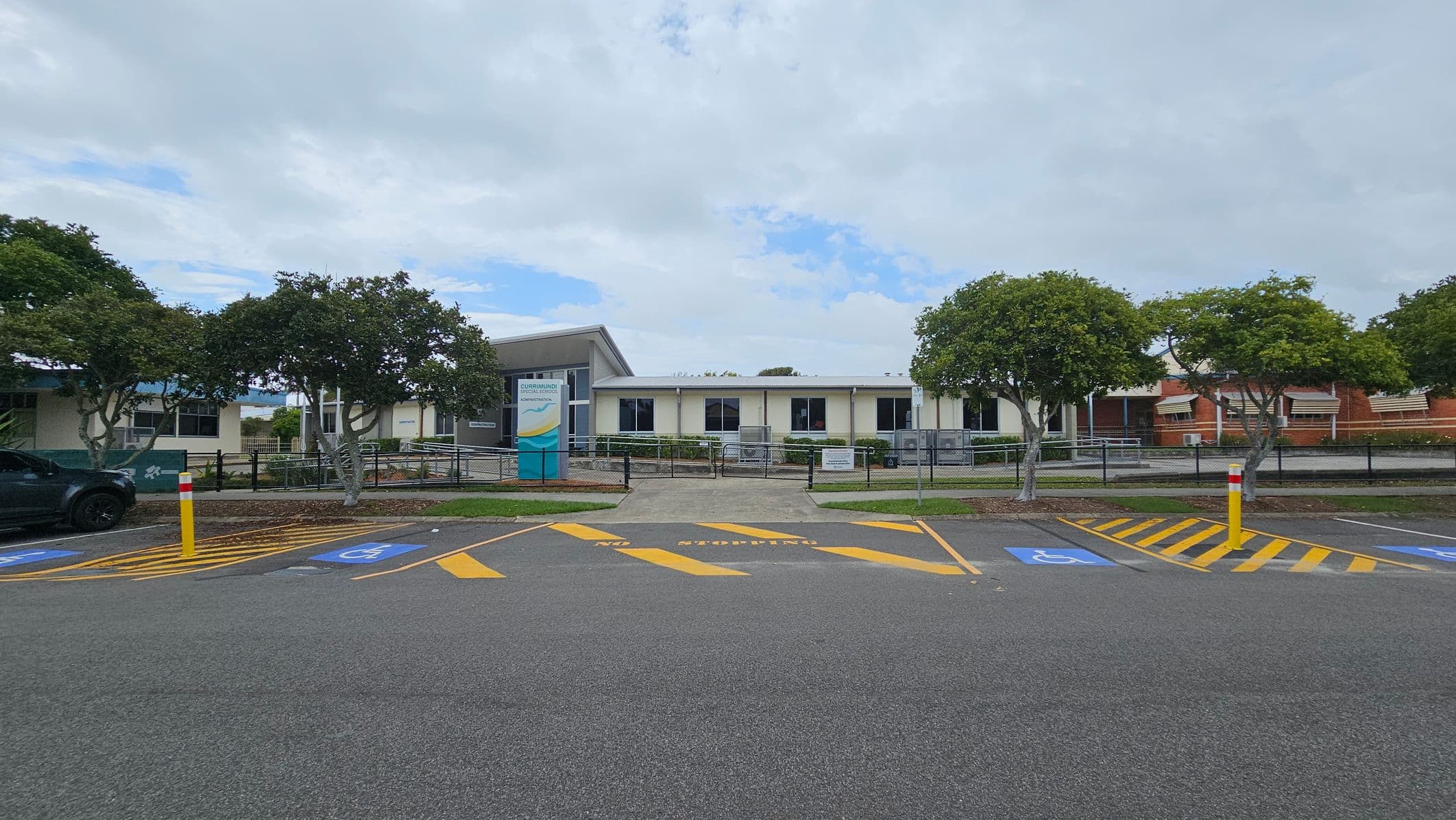 A single-storey building with a white facade and several windows is in the centre. In front, a sign with text is visible. The foreground features a car park with marked spaces, including disabled parking spots and yellow-striped areas. Trees with green foliage are on both sides, and a black fence runs along the front of the building. A grey asphalt surface covers the parking area.