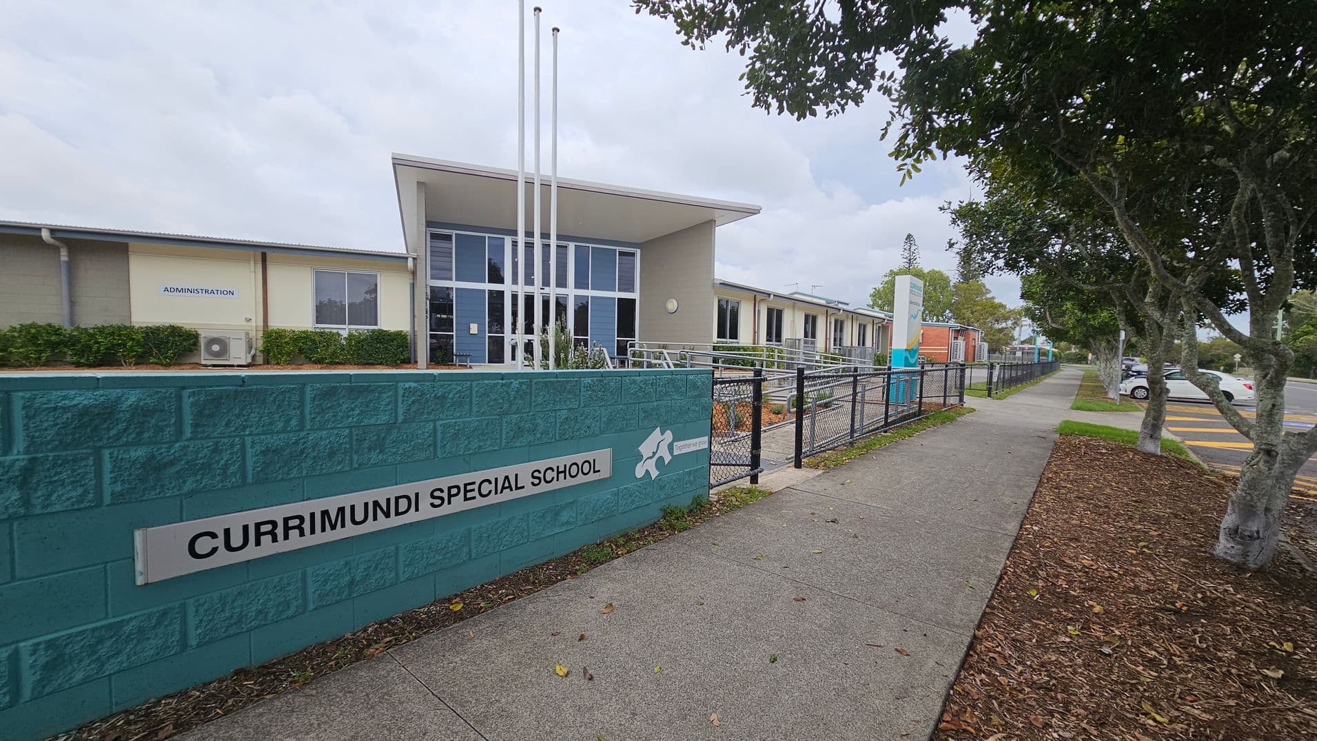 A school building with a modern design is on the left, featuring large windows and a sign reading "Administration." In front, a teal brick wall displays the name "Currimundi Special School." Three flagpoles stand nearby. On the right, a paved footpath runs alongside a fence, bordered by trees and a mulched area. Cars are parked in a lot further to the right.