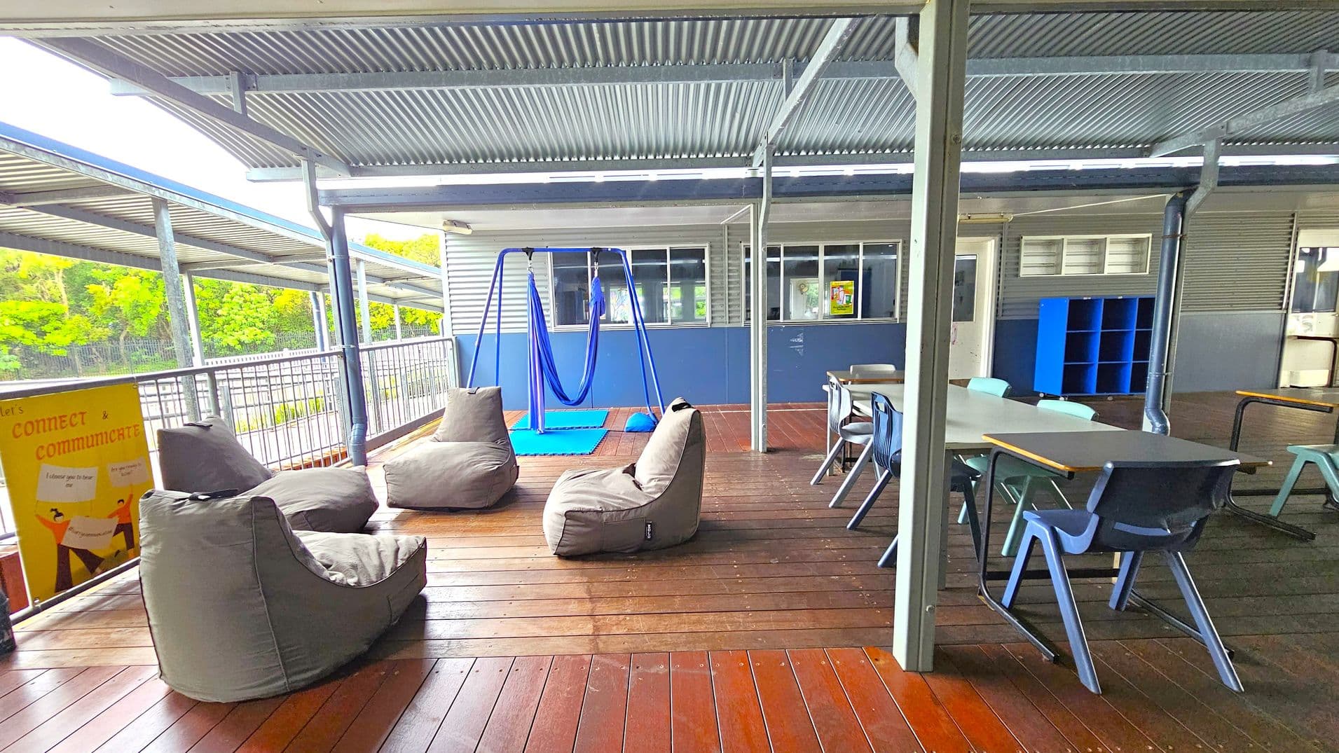 An outdoor covered area with a corrugated metal roof and wooden flooring. On the left, there are several grey bean bags and a yellow sign with text. In the centre, a blue swing hangs from a metal frame, with a blue mat underneath. On the right, there are tables and chairs, and a blue shelving unit against the wall. The background features a building with windows and a railing, with greenery visible beyond.