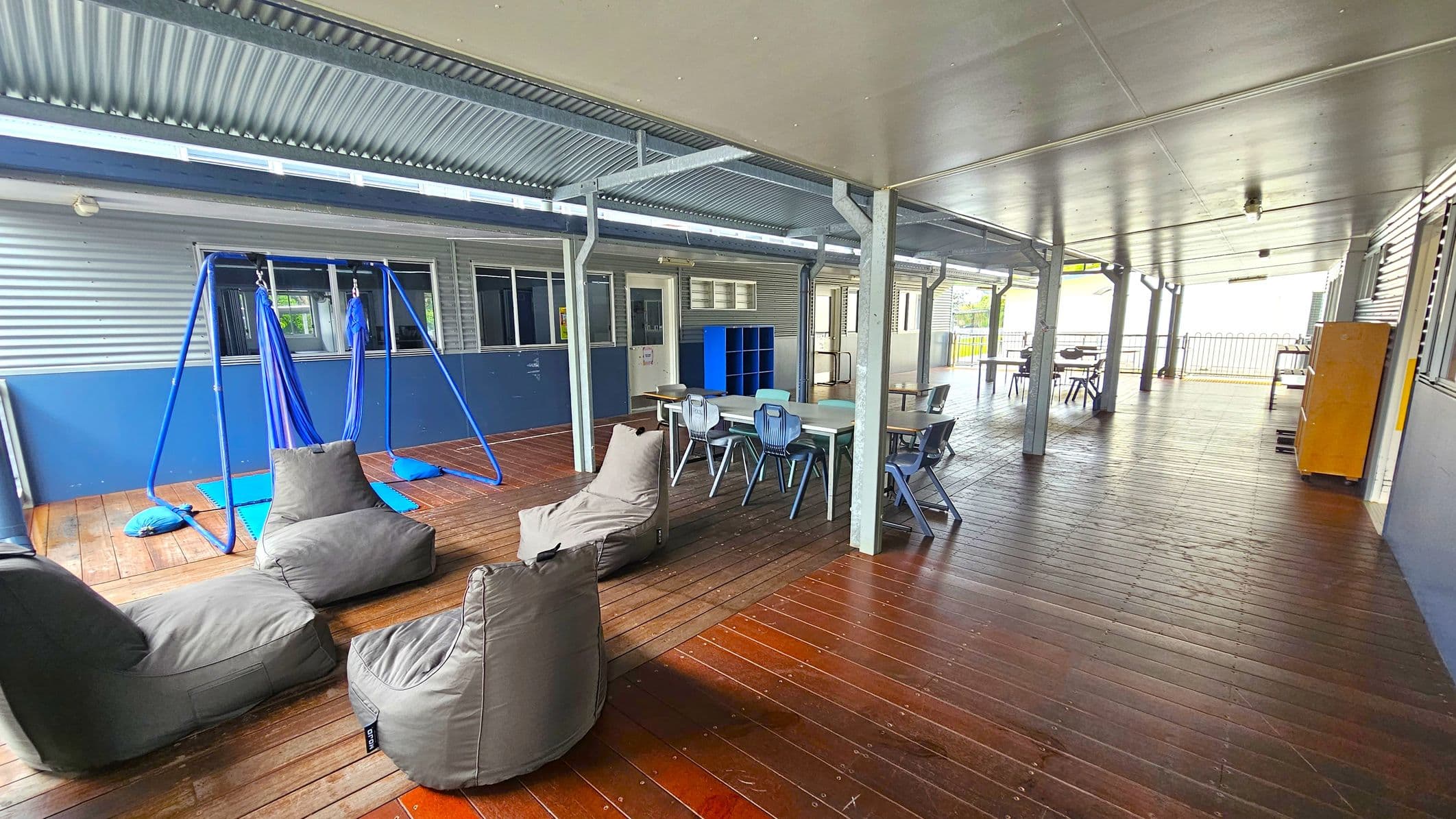 A covered outdoor area with a wooden floor and a metal ceiling. On the left, there are grey bean bags and a blue swing set with fabric seats. In the middle, a table with several blue chairs is positioned near a blue shelving unit. The right side features more tables and chairs, with a yellow cabinet against the wall. The background shows a corrugated metal wall with windows and doors, and a railing at the far end.