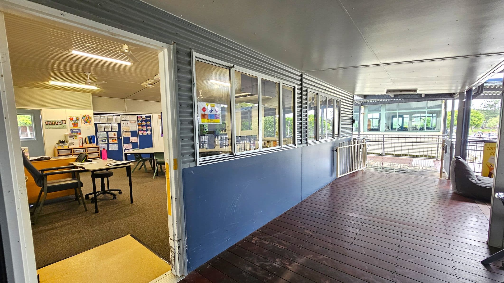 A corridor with a wooden floor and a metal ceiling. On the left, an open door leads to a classroom with a desk, chair, and educational posters on the walls. The classroom has a carpeted floor. The right side of the corridor features large windows with a blue lower wall. In the background, there is a railing and a view of another building with large windows and greenery visible outside.