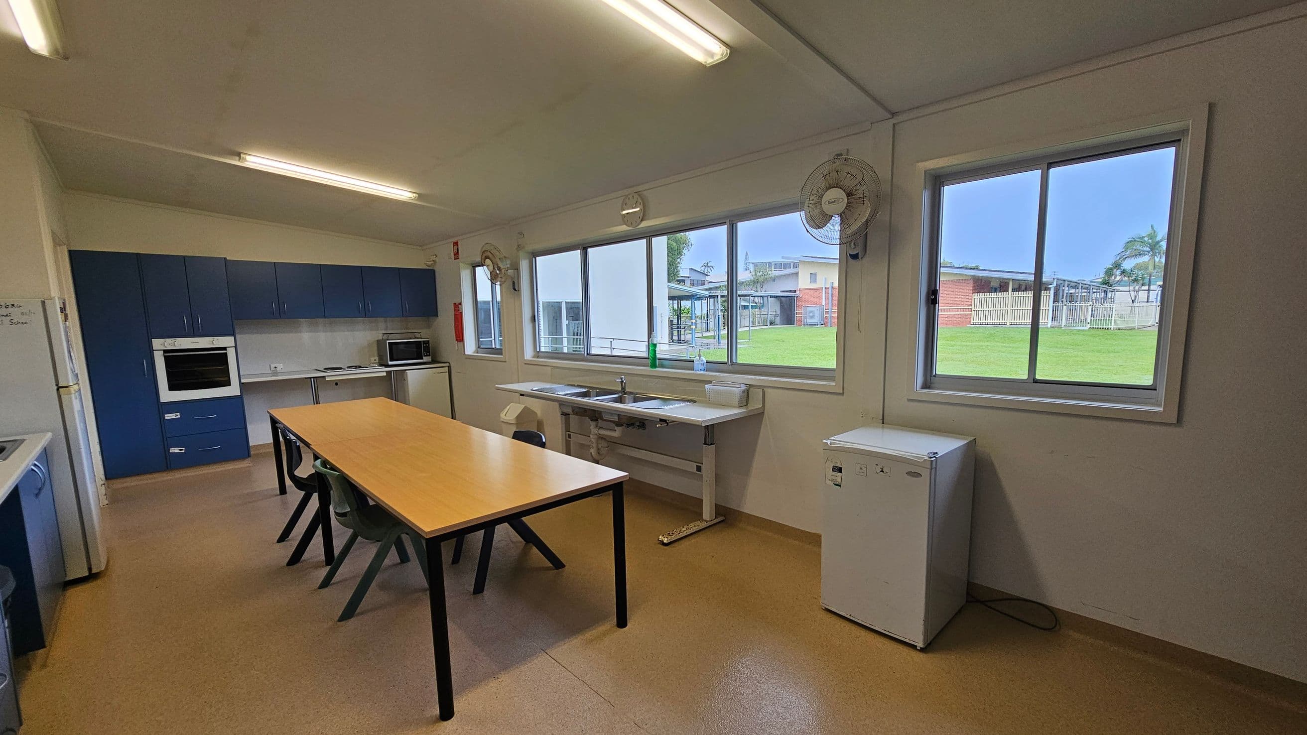 A kitchen with blue cabinets on the left, featuring an oven and a microwave. A long wooden table with black chairs is in the centre. Large windows on the right provide a view of a grassy area and buildings outside. Below the windows are two sinks and a small white fridge. The ceiling has fluorescent lights, and the floor is a light brown linoleum. Two wall-mounted fans are above the windows.