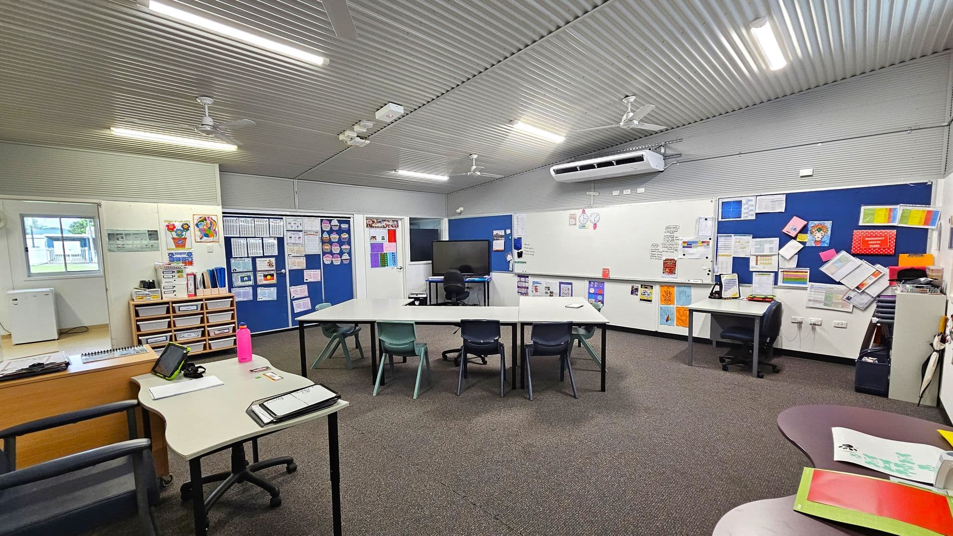 A classroom with a corrugated metal ceiling and fluorescent lights. The floor is carpeted. In the centre, there are tables arranged in a U-shape with various chairs. On the right, a large whiteboard and blue noticeboards are filled with papers and colourful materials. A desk with a chair and a monitor is also present. On the left, a window, storage drawers, and a small desk with a tablet and a pink water bottle. The walls are decorated with educational posters and charts.