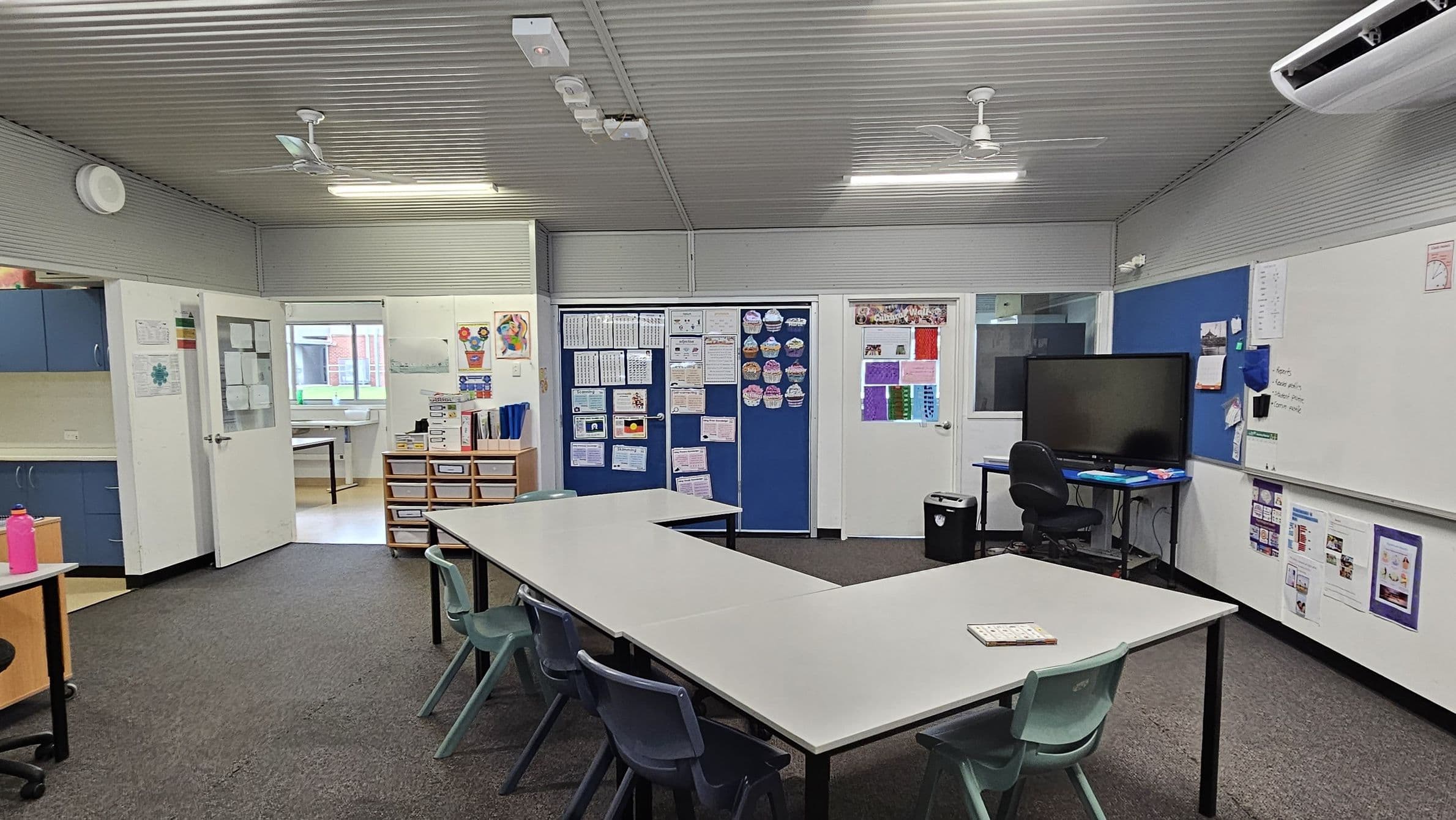 A classroom with a U-shaped arrangement of white tables and green chairs in the centre. On the right, there's a large whiteboard and a blue noticeboard with a flat-screen monitor on a desk. The back wall features blue and white doors with educational posters. On the left, blue cabinets and a pink water bottle are visible. The floor is carpeted, and the ceiling is corrugated with fluorescent lights and a ceiling fan. An air conditioning unit is mounted on the right wall.
