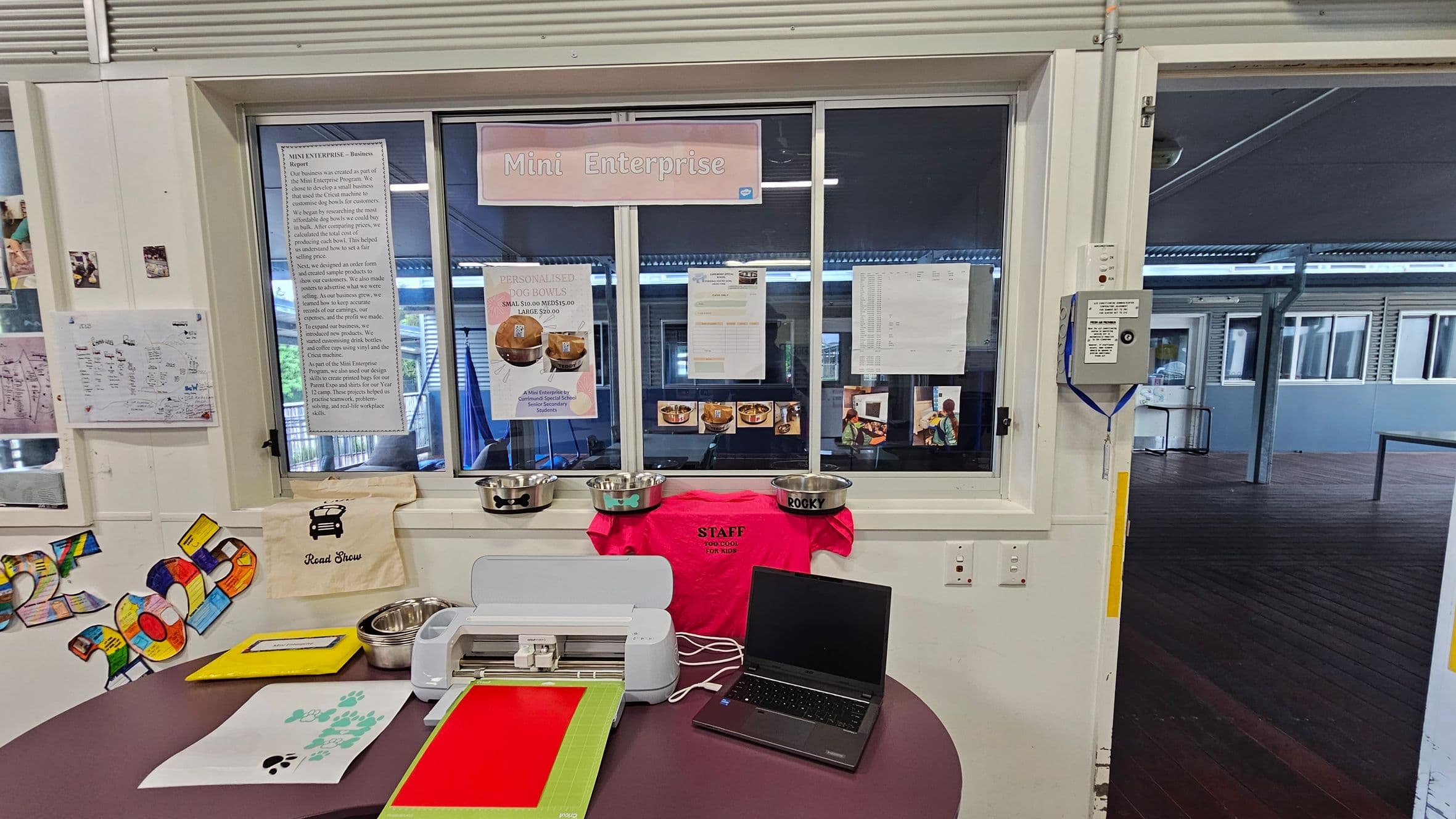 A room with a window labelled "Mini Enterprise" displaying various notices and posters. Below the window, there are several metal dog bowls, a bright pink staff T-shirt, and a laptop on a round table. The table also holds a cutting machine, colourful paper, and a sheet with paw prints. On the left wall, there are colourful cut-out numbers and a small poster. The floor is wooden, and the ceiling has visible beams. The background shows an open area with more windows and a wooden floor.
