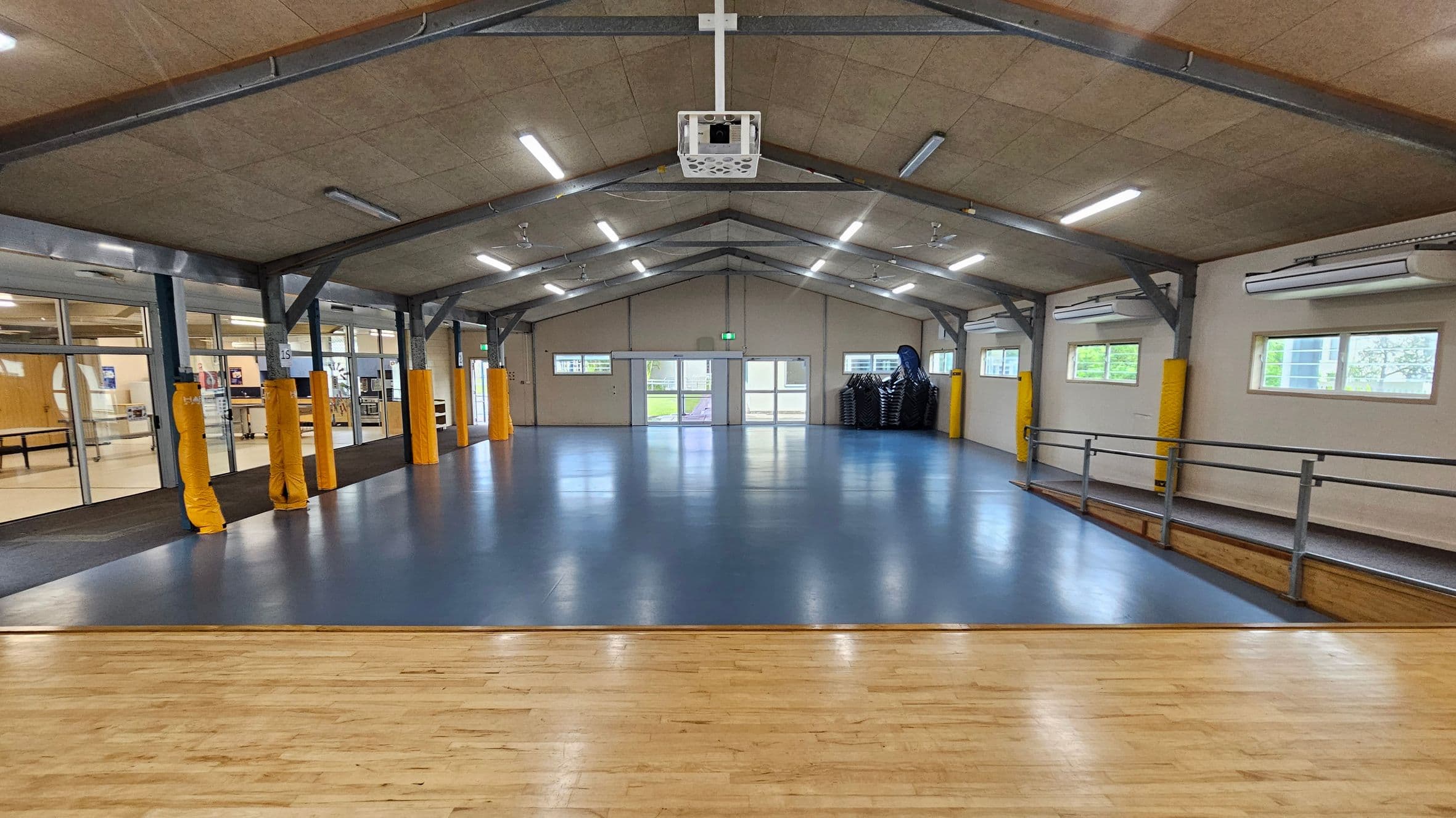 A spacious hall with a blue floor and a wooden stage in the foreground. The ceiling is high with exposed beams and fluorescent lights. On the left, there are large glass windows and doors leading to another room. On the right, windows allow natural light in, and a stack of chairs is visible. The back wall has double doors and small windows. Yellow padding covers some structural beams. The hall has a clean, open appearance.