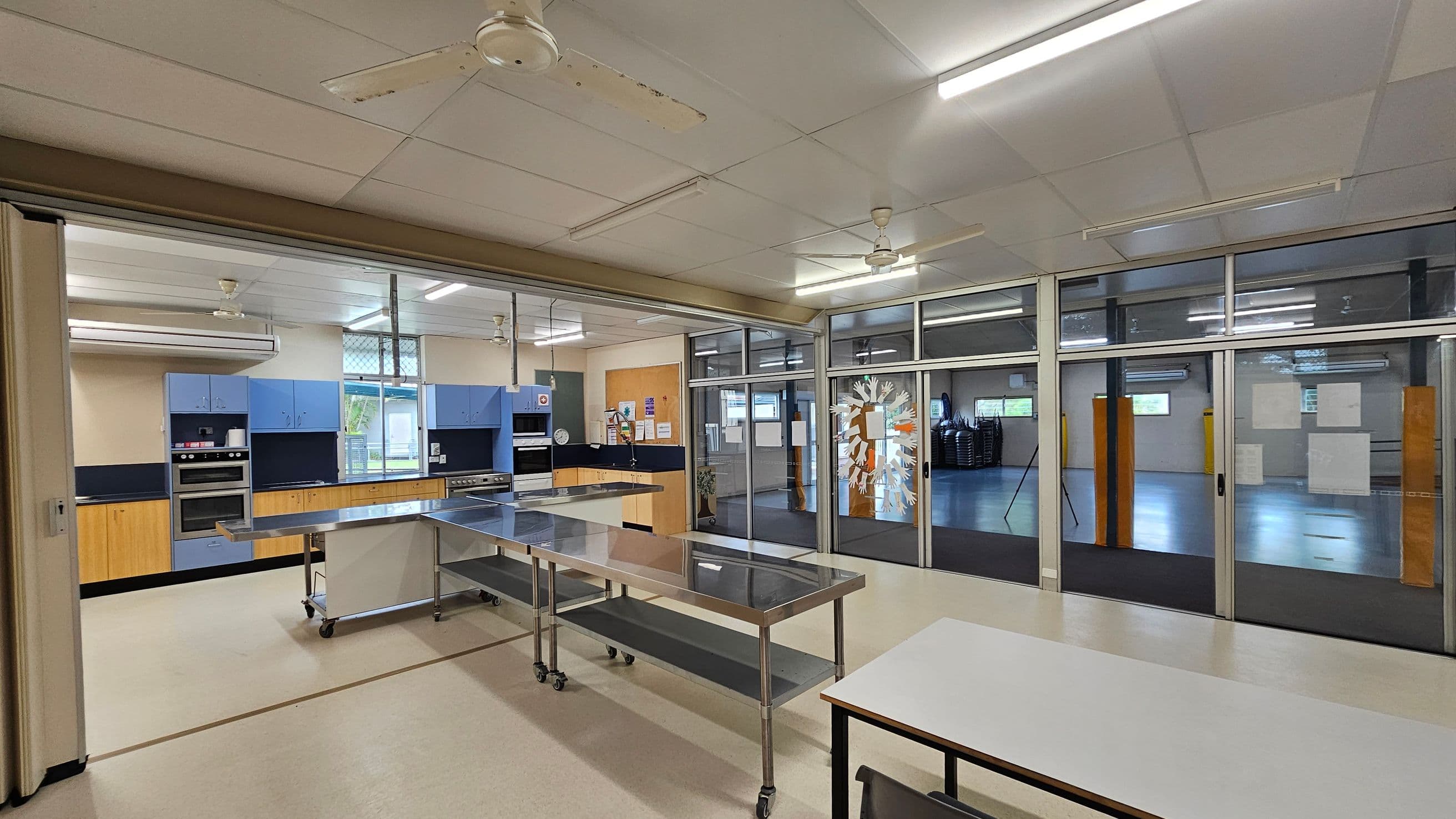 A spacious kitchen area with light-coloured flooring and a ceiling with fluorescent lights and ceiling fans. On the left, there are blue cabinets and a double oven. In the centre, a stainless steel table on wheels is positioned. The right side features large glass doors with paper decorations, leading to an adjacent room with stacked chairs and more windows. A white table is in the foreground.