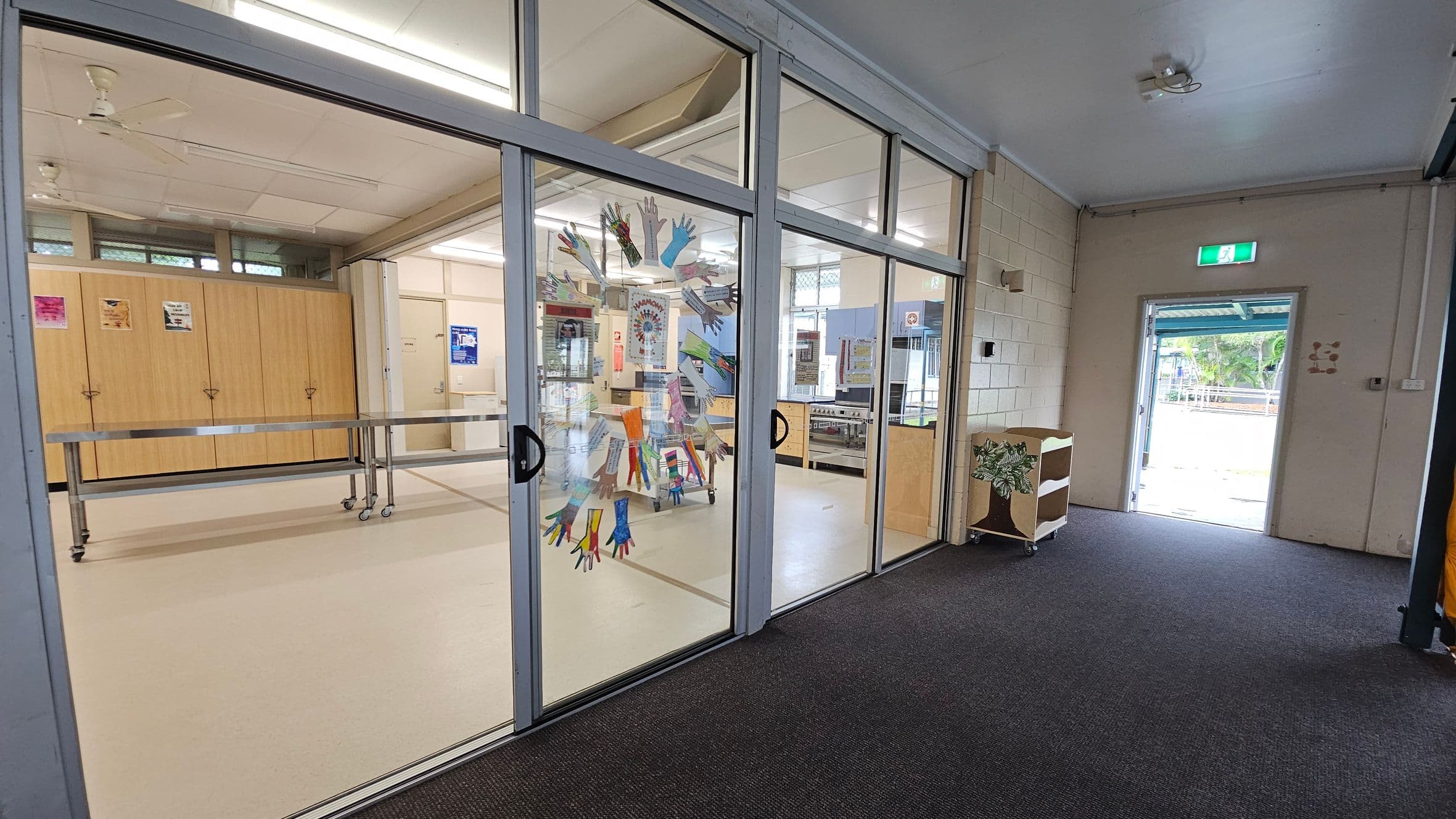 A room with glass sliding doors decorated with colourful handprints. Inside, there are wooden cabinets and a metal table on wheels. The ceiling has fluorescent lights and a ceiling fan. The floor is carpeted in the foreground and tiled inside the room. To the right, an open door leads outside, with a small shelf nearby. Posters are visible on the walls and cabinets.