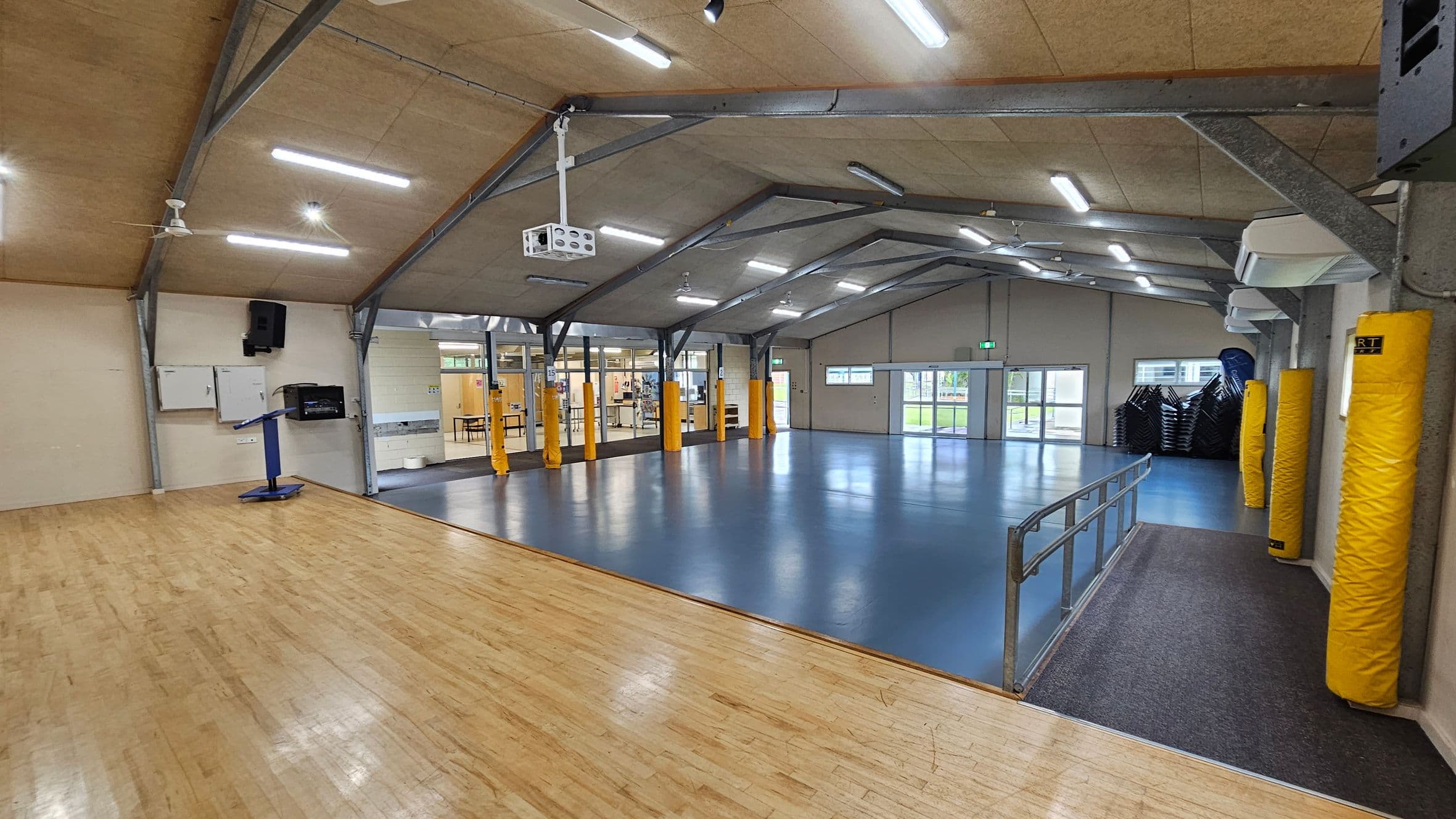 A spacious indoor hall with a wooden floor on the left and a blue floor in the centre and right. The ceiling is high with exposed beams and fluorescent lights. Yellow padded columns are evenly spaced along the walls. A projector is mounted on the ceiling. On the right, there are stacked chairs near a set of double doors. A metal railing separates the wooden and blue floor areas. The background features glass doors and windows leading to another room.