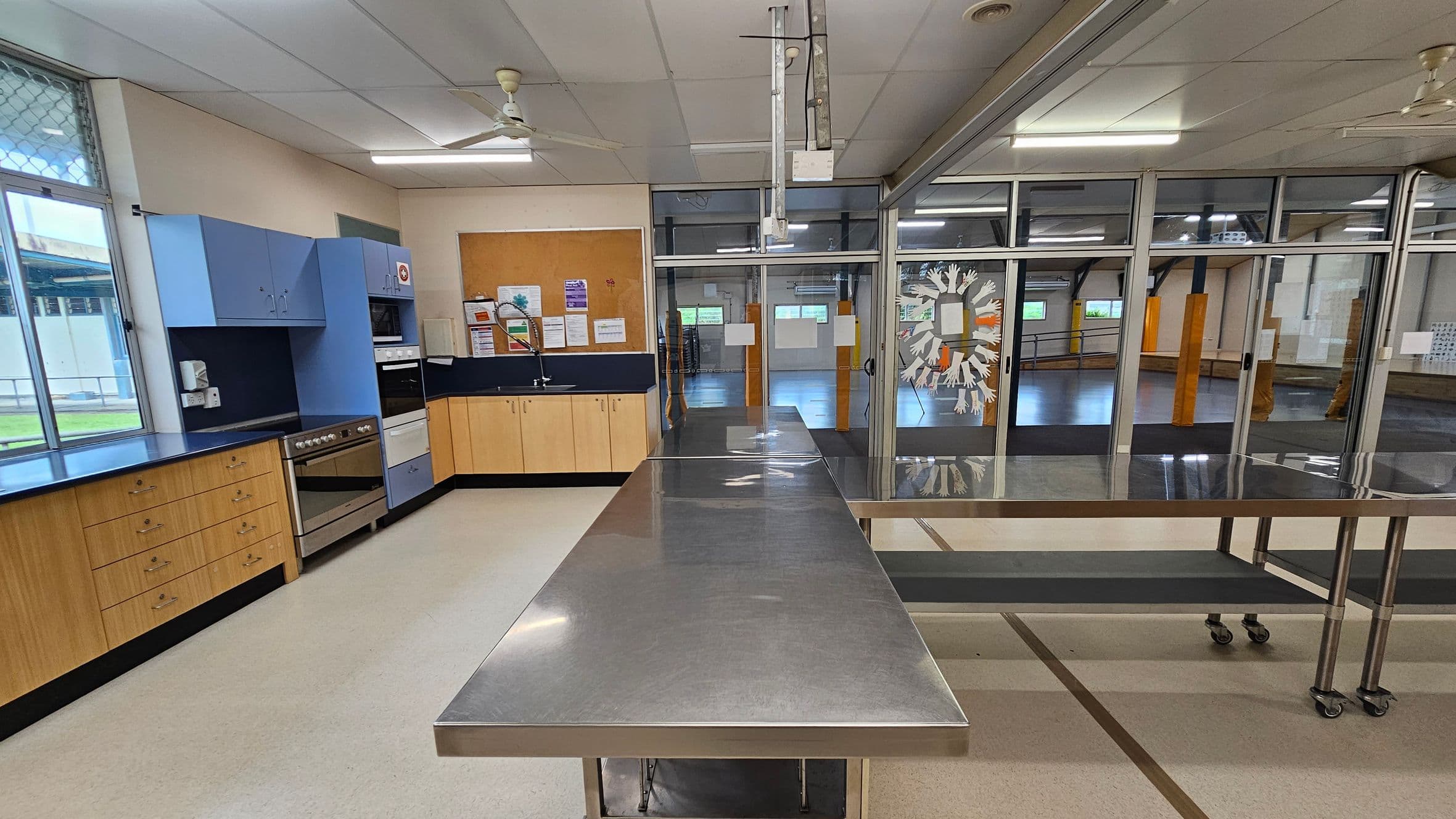 A kitchen with light-coloured flooring and a white ceiling with fluorescent lights and ceiling fans. On the left, there are wooden cabinets with a blue countertop, a stainless steel oven, and a microwave. A window with a mesh screen is above the counter. In the centre, a long stainless steel table on wheels. The right side features glass doors with a decorative design, leading to a large open room with visible support beams and windows.