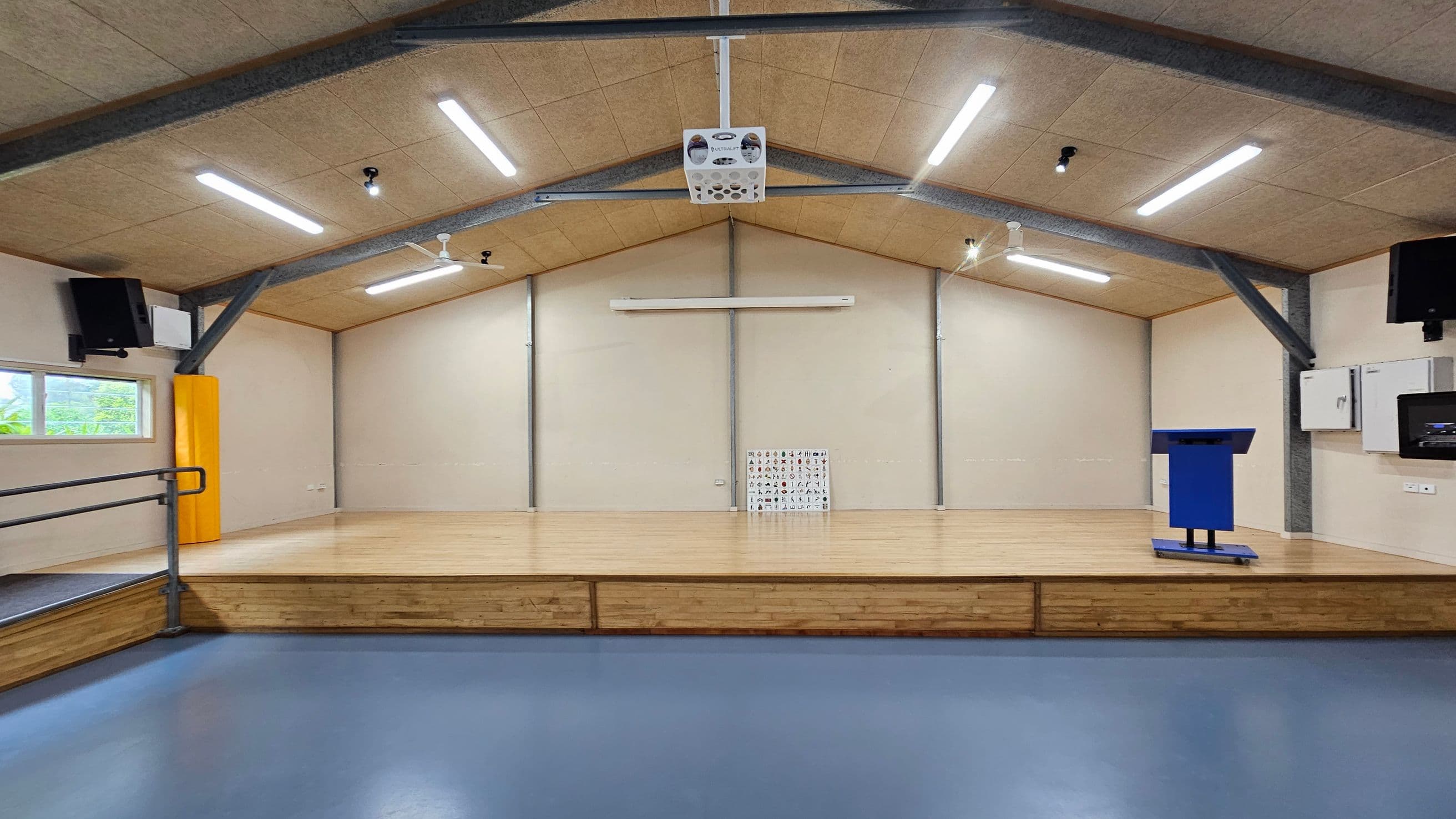 A spacious room with a wooden stage in the centre, featuring a blue podium on the right. The floor is blue, and the ceiling has exposed beams with several fluorescent lights. On the left, there's a window with a yellow padded column nearby. The back wall has a small chart with various symbols or letters. Two black speakers are mounted on either side of the room.