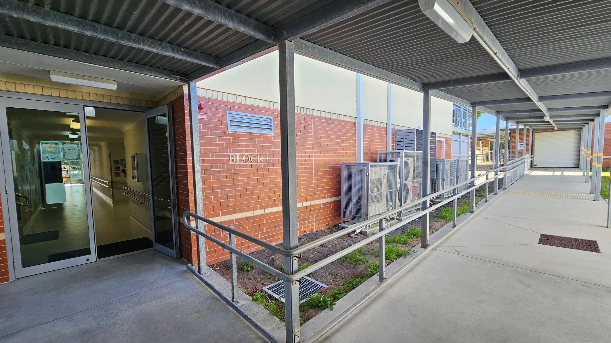 A covered walkway with a metal roof and railings leads to a building entrance on the left. The entrance has glass doors, and inside, a hallway is visible. The building is made of red brick with a sign reading "BLOCK C." Air conditioning units are mounted on the exterior wall. The floor is concrete, and there is a small patch of grass and soil beside the walkway. The ceiling has fluorescent lights.