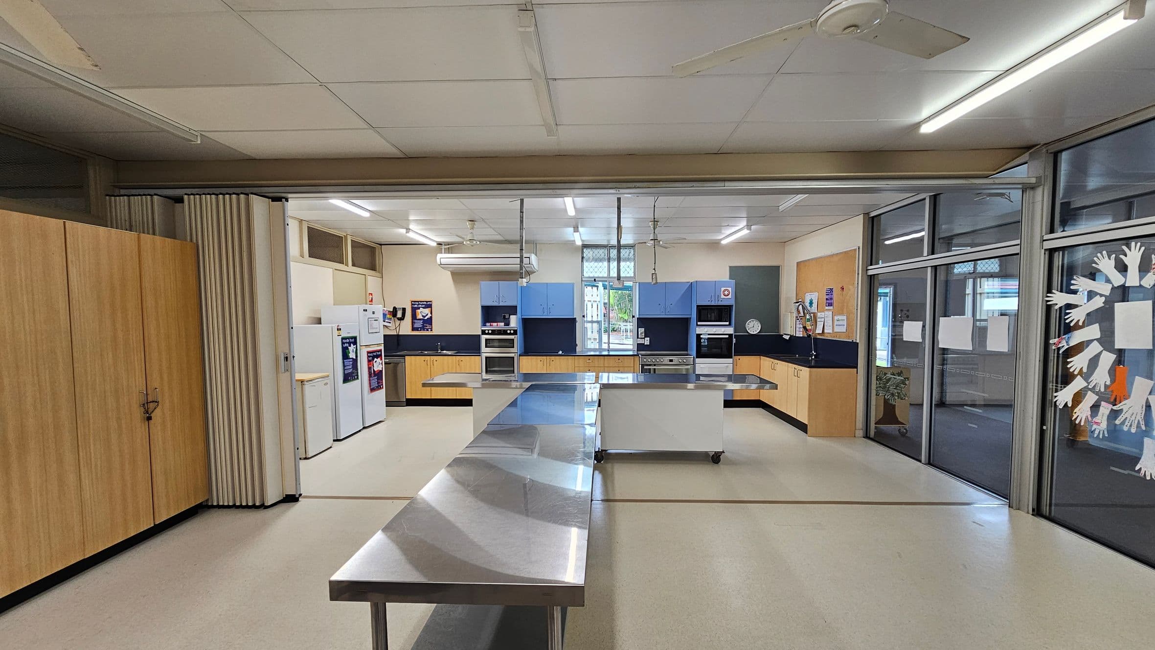 A spacious kitchen with a central stainless steel workbench. On the left, there are wooden cabinets and a row of white appliances, including fridges. The back wall features blue and wooden cabinetry with built-in ovens and a sink. On the right, large glass windows display paper cut-outs of hands. The ceiling has fluorescent lights and a fan, and the floor is a light-coloured linoleum.