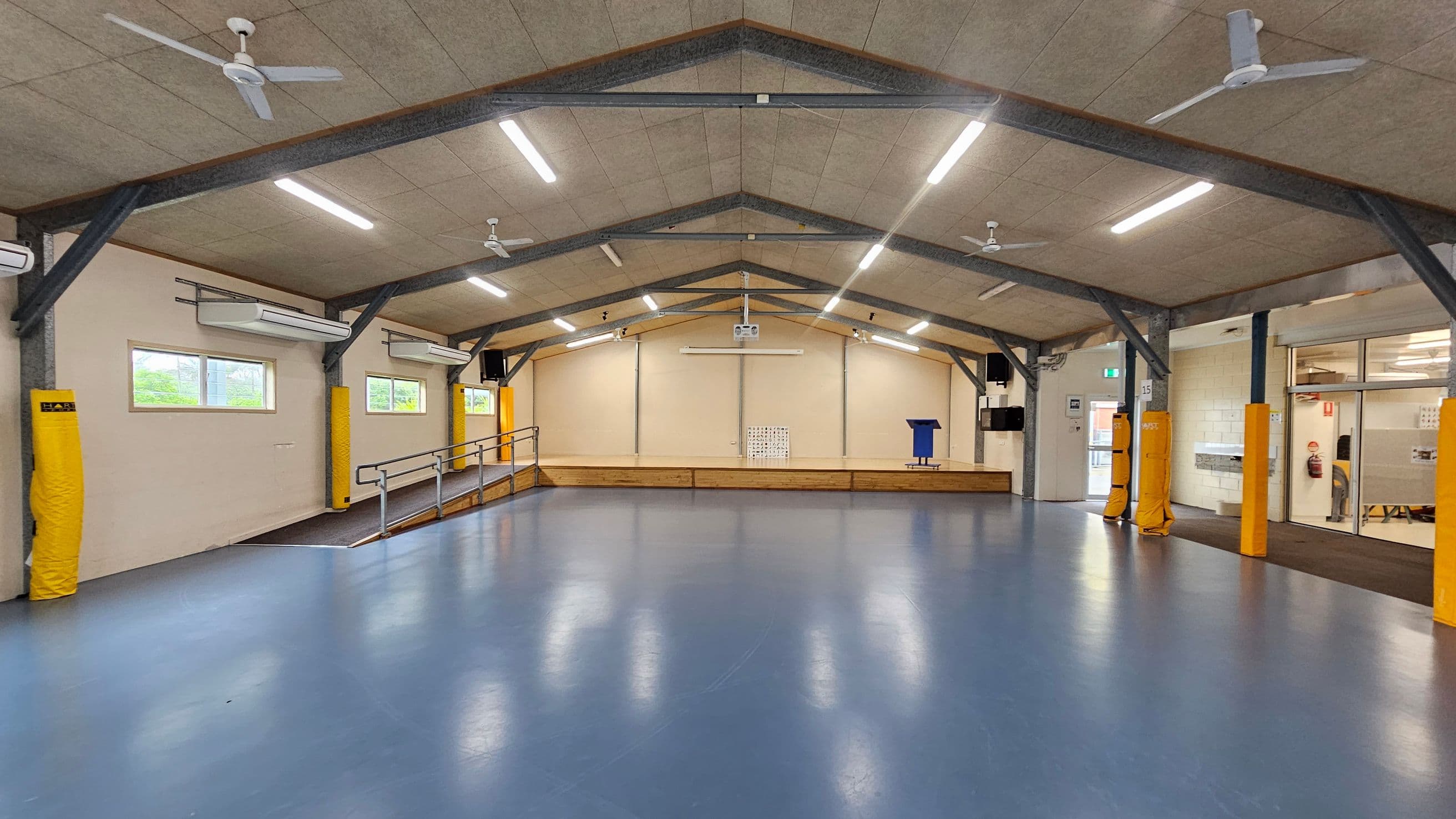 A spacious indoor hall with a blue floor and a high, sloped ceiling featuring exposed beams and ceiling fans. The left side has windows with air conditioning units above, and yellow padding on the beams. The right side shows a glass entrance with a door and a fire extinguisher nearby. In the centre, a stage with a ramp, a blue lectern, and a projector screen is visible. The ceiling has multiple fluorescent lights.