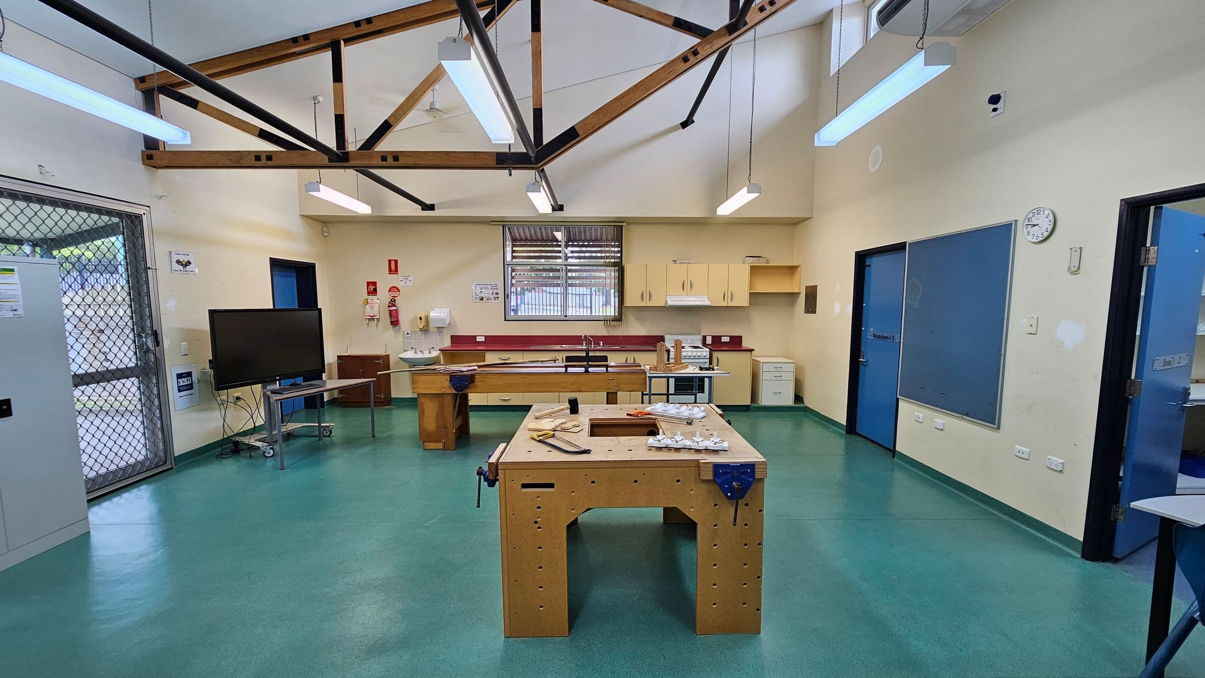 A workshop room with a green floor and high ceiling featuring exposed wooden beams. In the centre, a wooden workbench is equipped with tools and materials. The back wall has a window with a metal grille, surrounded by cabinets and a sink. On the left, a large TV on a stand is near a metal mesh door. The right side has blue doors and a clock on the wall. Fluorescent lights hang from the ceiling.