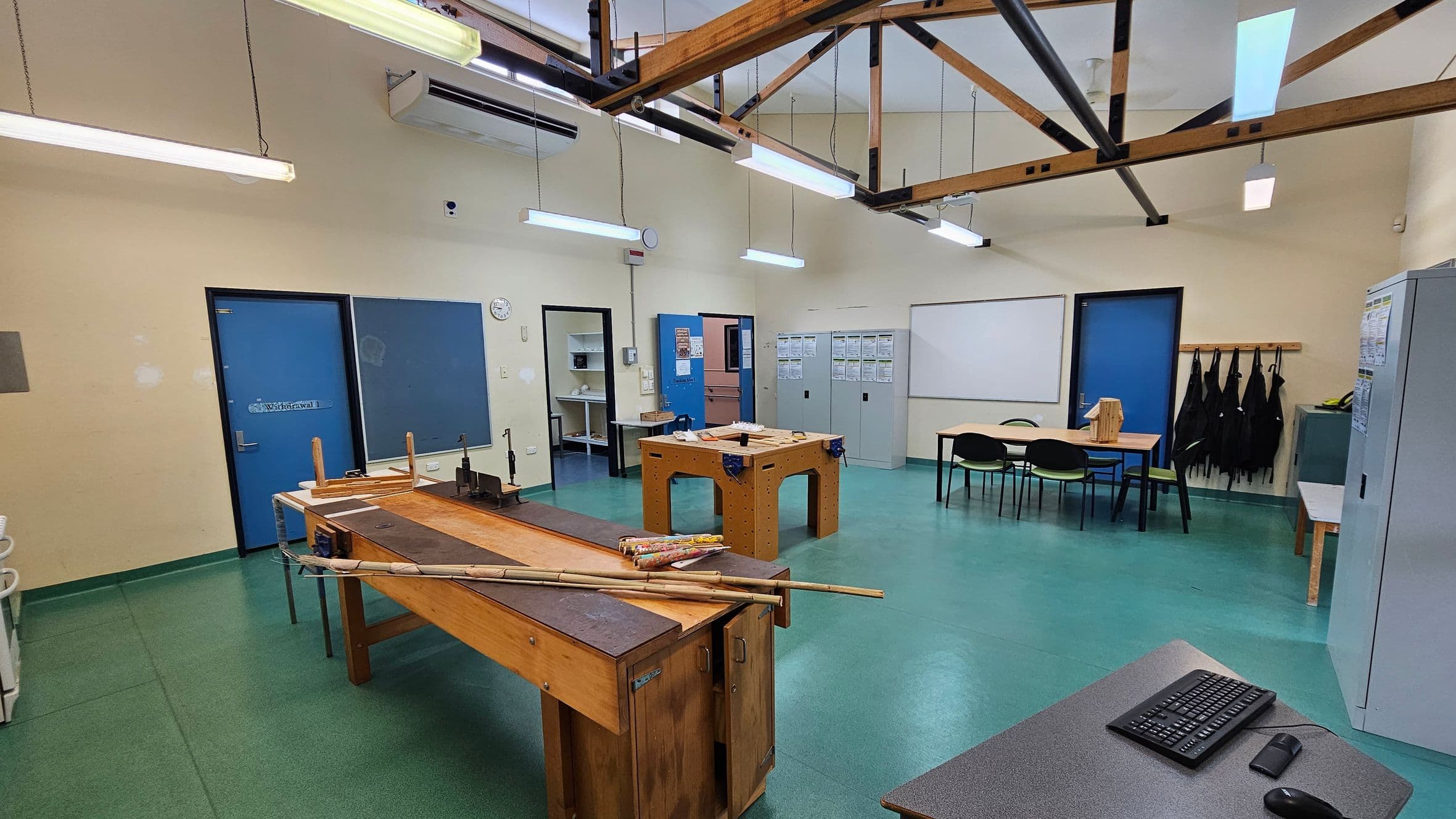 A workshop room with green flooring and a high ceiling featuring exposed wooden beams. In the foreground, a wooden workbench with tools and materials is visible. To the right, a grey table with a keyboard and mouse. The background has blue doors, a clock, and a whiteboard. Lockers and a table with chairs are on the right, with black aprons hanging on hooks. Fluorescent lights hang from the ceiling.