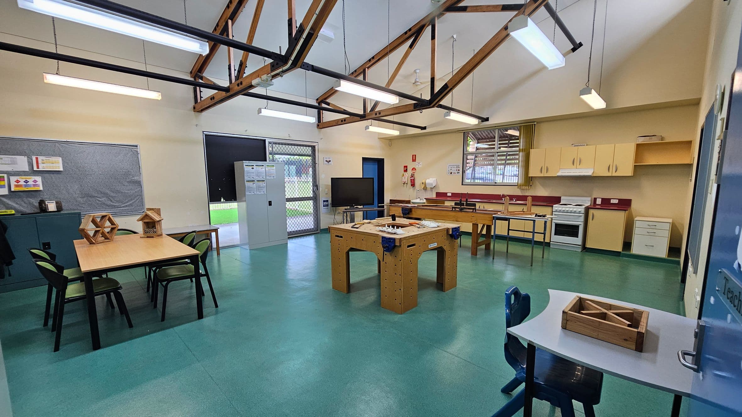 A classroom with a green floor and high ceiling featuring exposed wooden beams. On the left, there's a table with chairs and geometric wooden models. A bulletin board and storage cabinet are nearby. In the centre, a large wooden workbench is visible. The right side has a kitchen area with cabinets, a stove, and a sink. A small desk with a chair and wooden box is in the foreground. Fluorescent lights hang from the ceiling, and a door and windows are in the background.