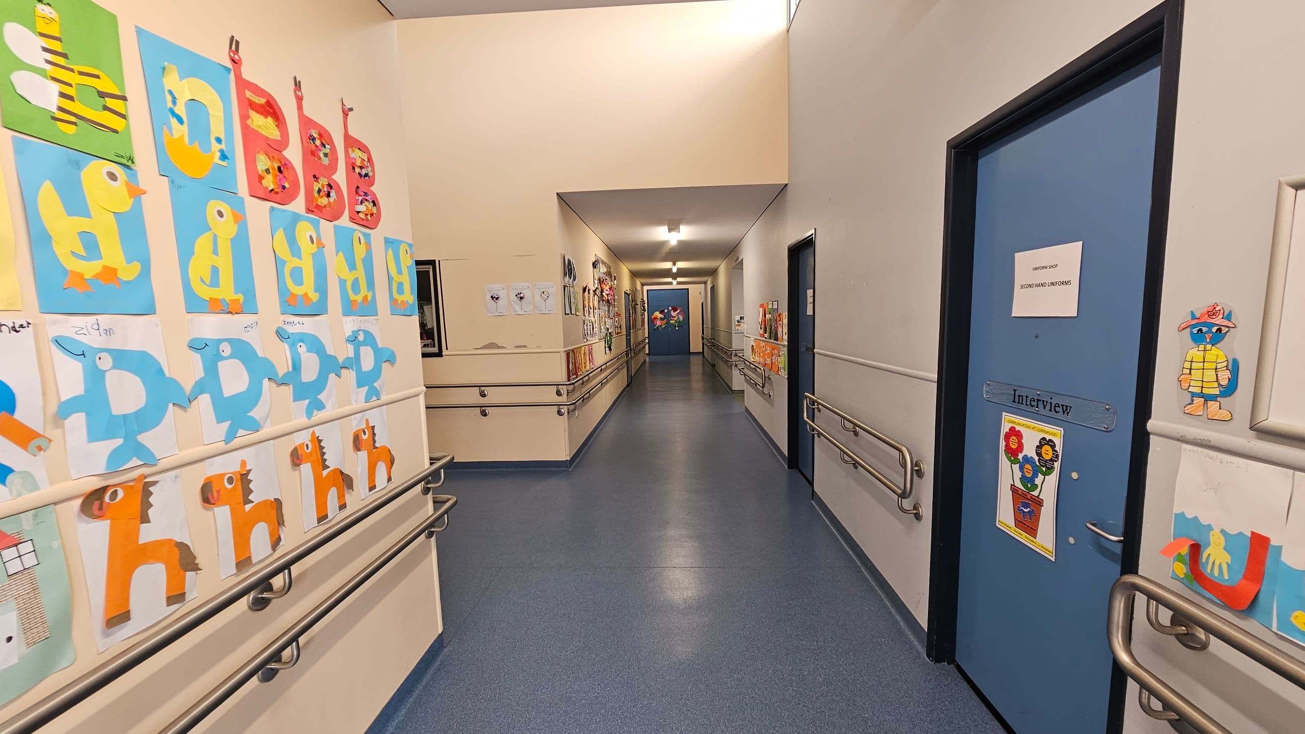 A corridor with blue flooring and beige walls. On the left, colourful children's artwork featuring animals and letters is displayed. On the right, a blue door with a sign reading "Interview" and more artwork is visible. Handrails run along both sides of the corridor. The ceiling is white with fluorescent lighting, and more artwork is visible further down the hallway.