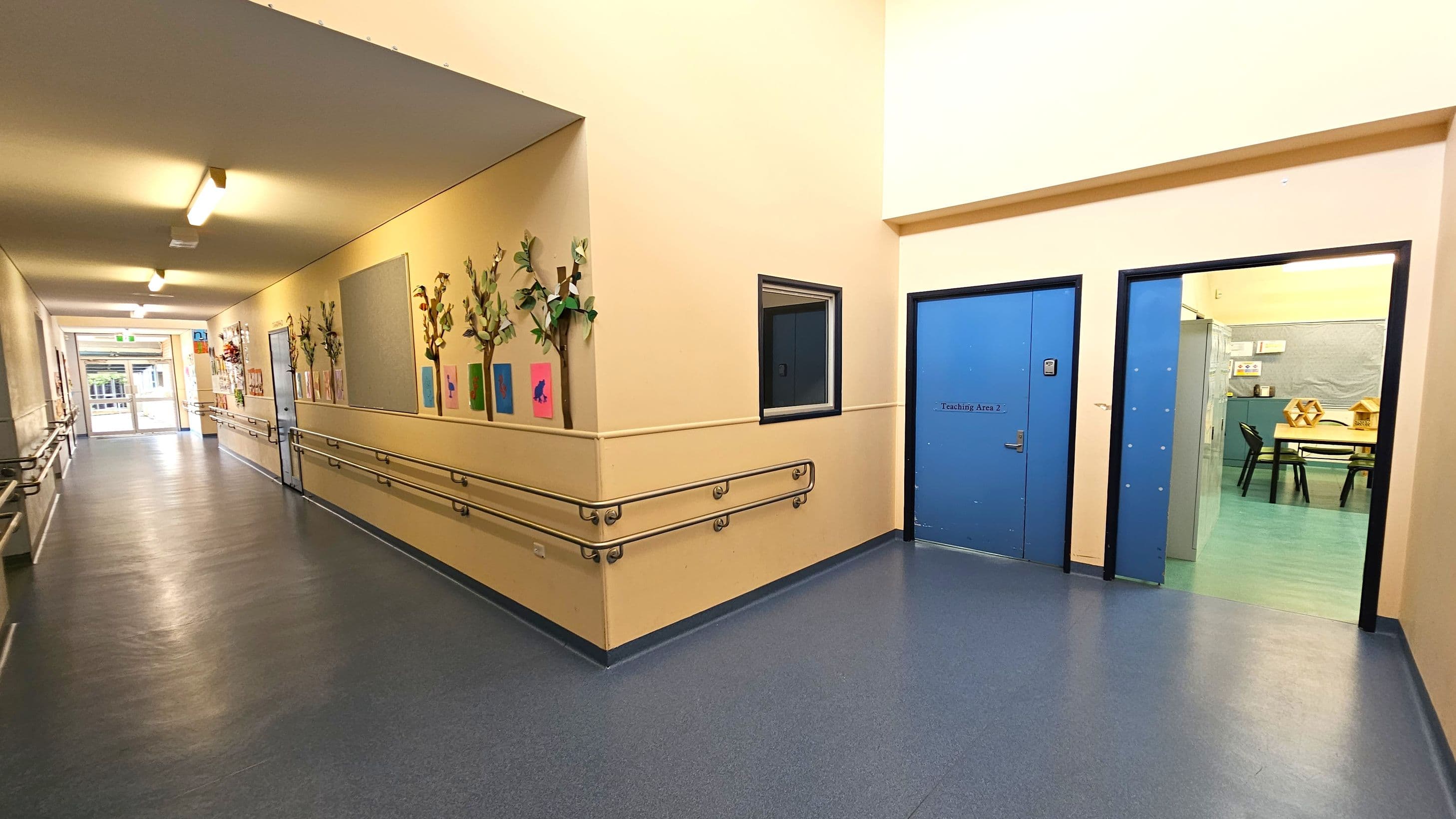 A corridor with a blue floor and cream walls. On the left, the wall is adorned with colourful artwork and decorative plants. Handrails run along both sides. At the end of the corridor, there is a glass door leading outside. On the right, there are two blue doors, one labelled "Teaching Area 2," and an open doorway revealing a room with tables and chairs. The ceiling is cream with fluorescent lights.