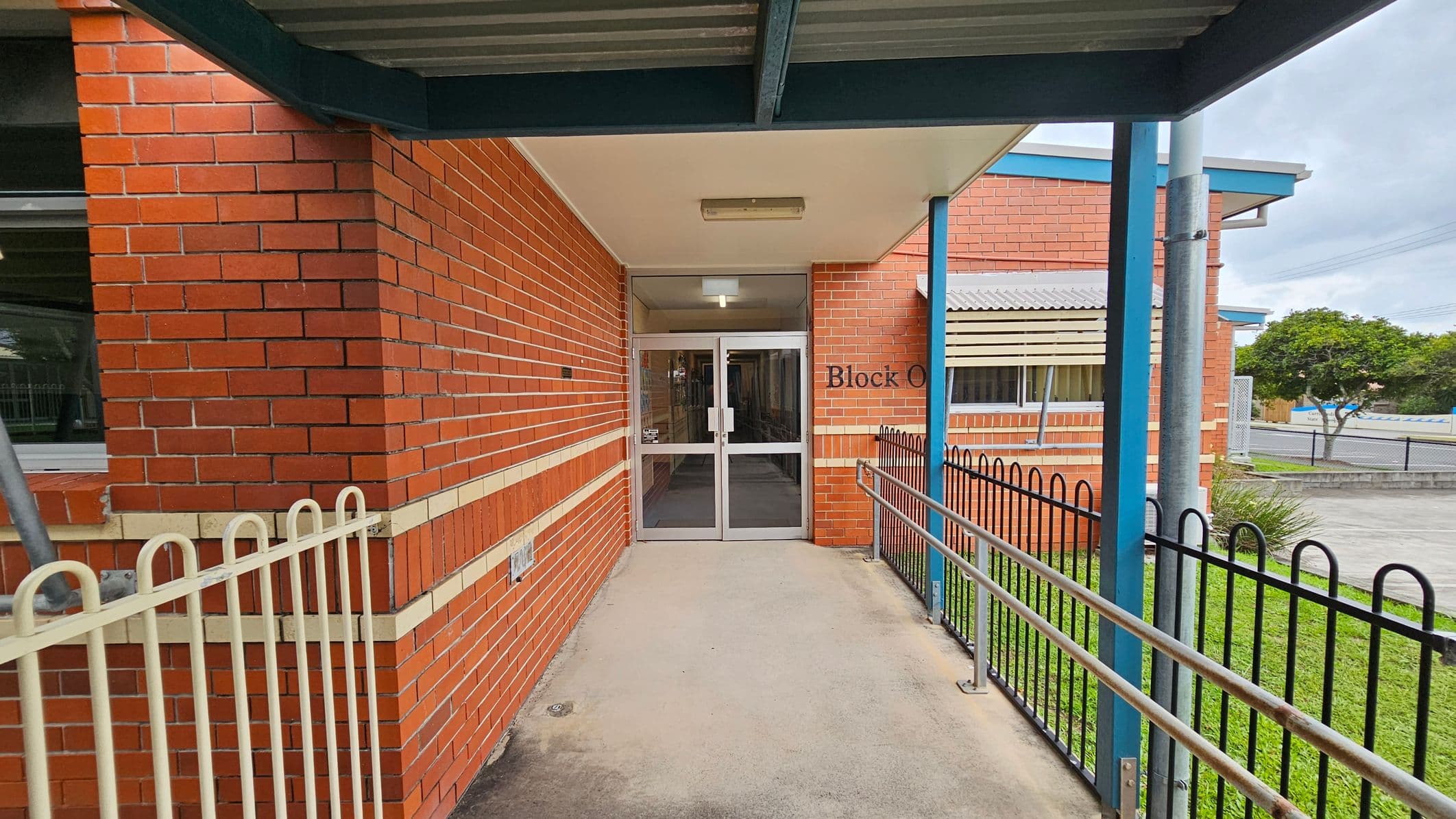 A brick building entrance with a sign reading "Block O" above the glass double doors. The left side features a red brick wall with a horizontal beige stripe and a window. On the right, there's a black metal fence and a grassy area. A blue metal post supports the overhanging roof. The concrete pathway leads to the entrance, and the ceiling is plain with a light fixture. In the background, there's a tree and a paved area.