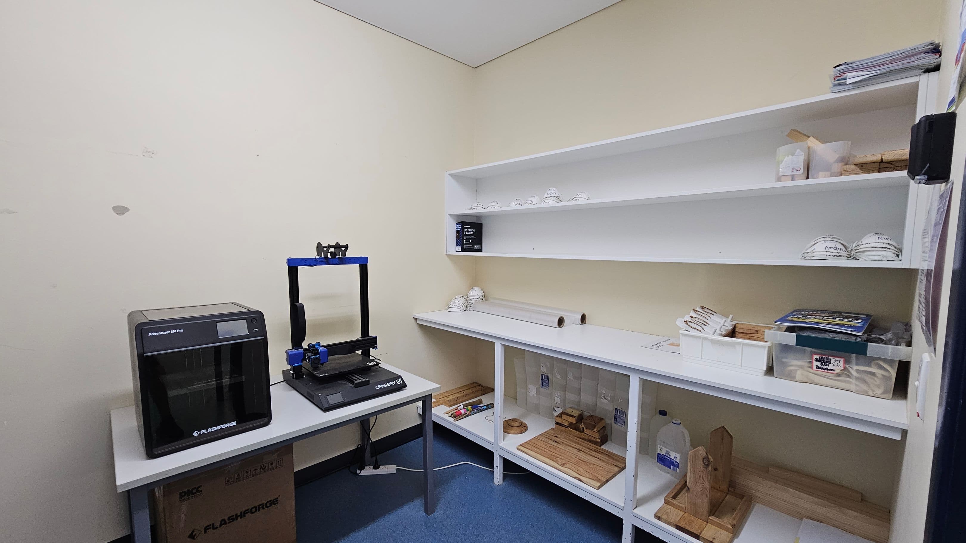A small room with a blue floor and a white ceiling. On the left, there is a 3D printer on a table, with a cardboard box underneath. In the middle, white shelves hold various items, including stacked white objects, a box, and rolls of material. On the right, more shelves contain trays, books, and wooden items. The walls are a light colour, and the room appears organised for storage or work.