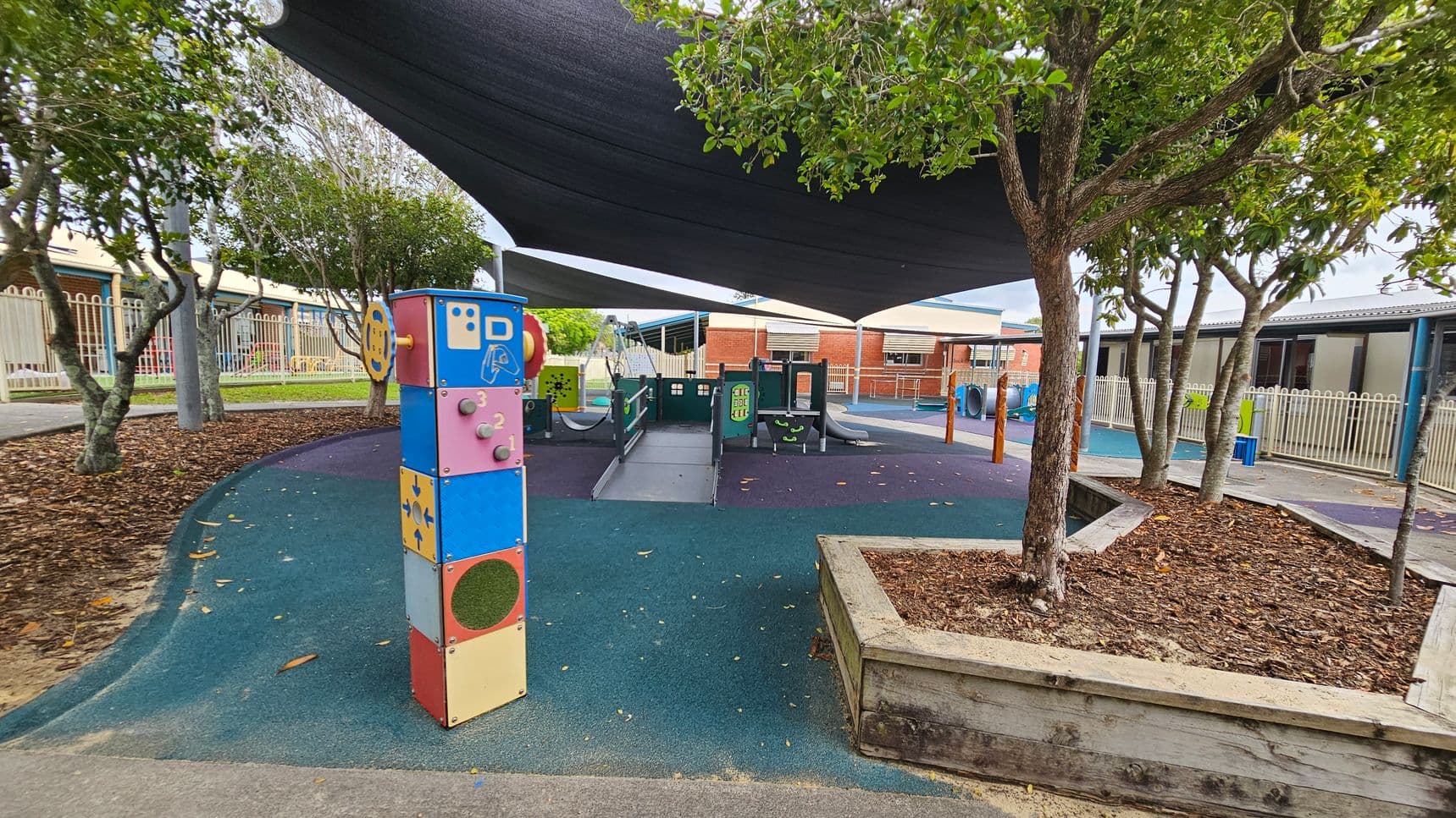 A playground with a colourful interactive panel on the left, featuring various shapes and numbers. In the centre, there's a play structure with slides and climbing elements. The ground is covered with a blue and green rubber surface. Trees with green foliage are on the right, and a wooden planter box is visible. Overhead, a large black shade cloth provides cover. In the background, there are buildings with brick and metal exteriors, and a white fence runs along the perimeter.
