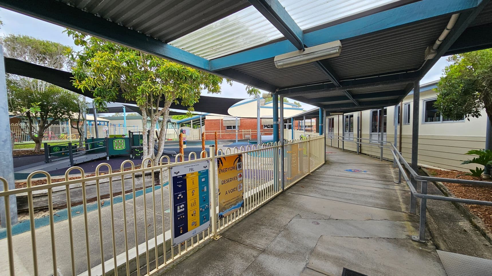 A covered walkway with a corrugated metal roof and transparent panels runs alongside a building with multiple windows on the right. A metal railing lines the path, with signs attached. On the left, a fenced playground area features trees, play equipment, and shade structures. The ground is a mix of concrete and rubberised surfaces. In the background, brick buildings are visible, and the area is surrounded by more fencing and greenery.