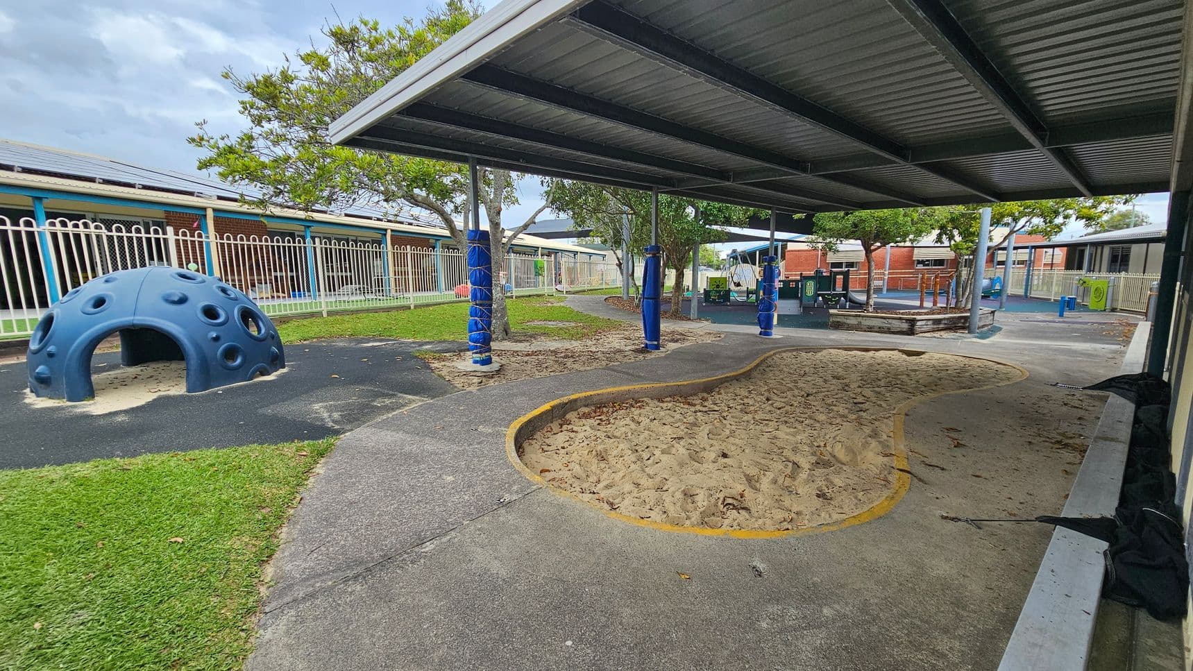 A playground with a blue dome-shaped climbing structure on the left, surrounded by grass and a paved area. In the centre, there's a sandpit with a yellow border. The right side features a covered area with a metal roof supported by poles wrapped in blue padding. In the background, there are trees, a fence, and school buildings with blue and red accents. The ground is a mix of grass, sand, and pavement.