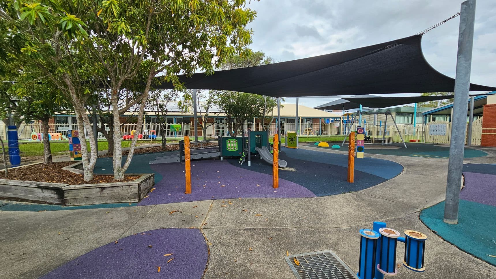 A playground with a variety of equipment, including a slide and interactive panels, is covered by large black shade sails. The ground is a mix of purple and blue soft surfaces. On the left, there are trees in a raised wooden planter. In the background, a fence encloses the area, with a building visible beyond it. The foreground features a concrete path and a small blue musical instrument.