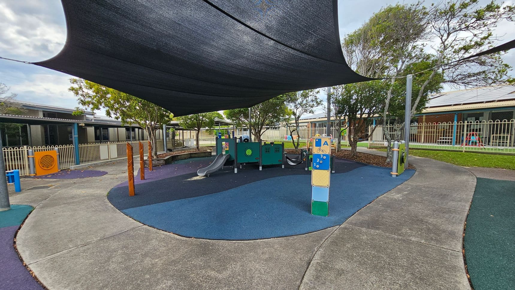 A playground with a blue and green rubber surface. In the centre, there is a play structure with slides and climbing features. A large black shade sail is overhead. Trees and a fence line the background, with a building visible behind them. The ground is a mix of blue and green pathways, and there are wooden posts and interactive play panels scattered around.