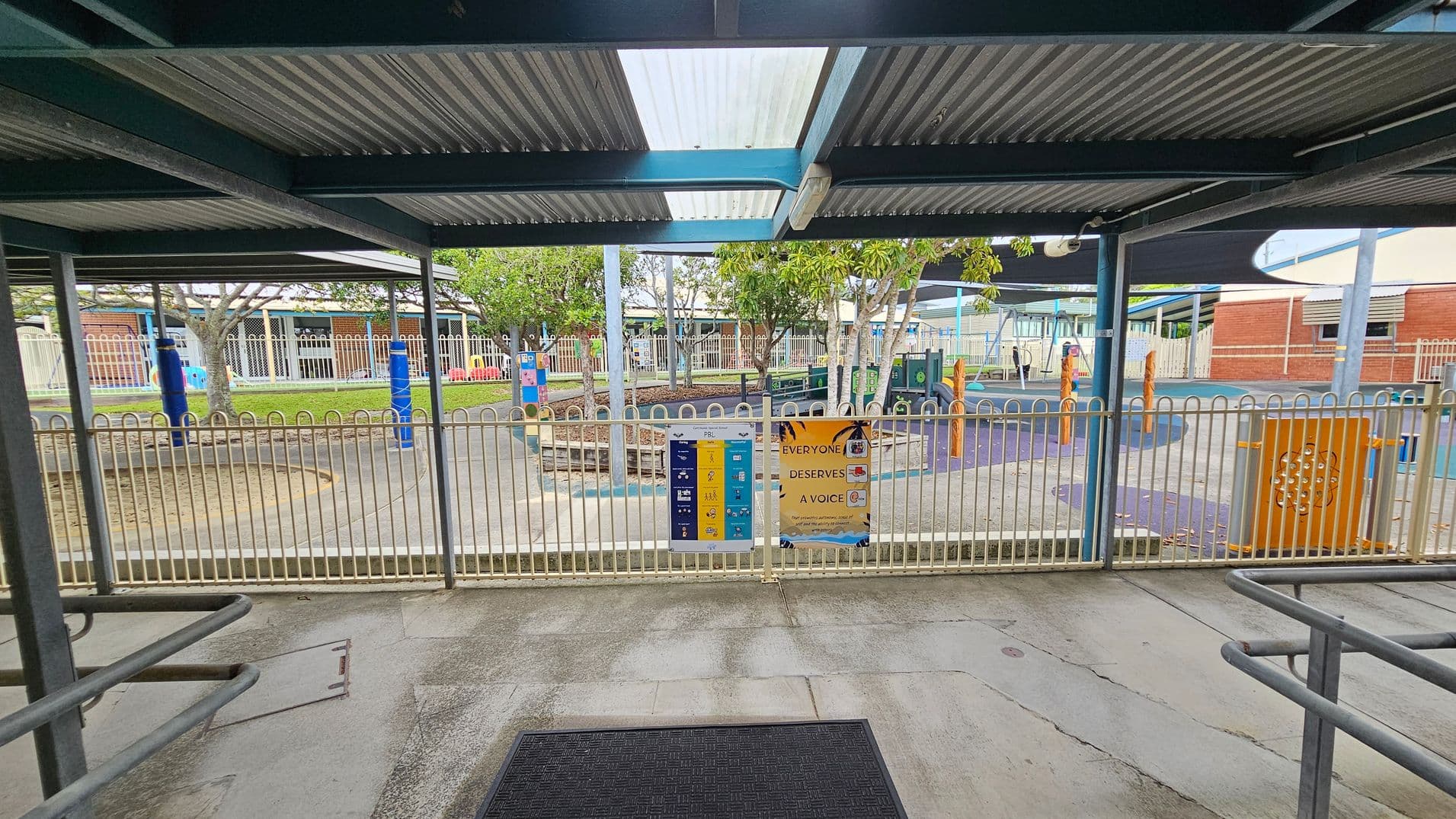 A covered outdoor area with a corrugated metal roof and a black mat on the concrete floor. Beyond the metal fence, there is a playground with colourful equipment, including blue and orange structures. Trees provide some shade, and there are signs on the fence with text and graphics. In the background, there are buildings with brick and metal elements, and a shaded area with a dark canopy.