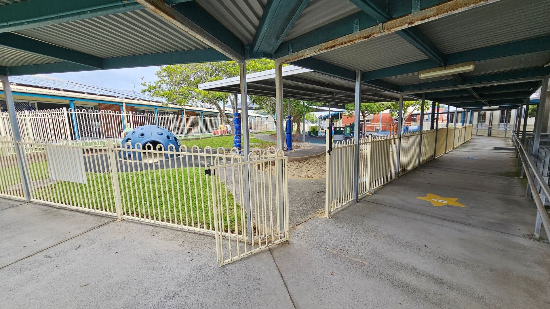 A covered walkway with a corrugated metal roof and metal support beams. On the left, there's a fenced playground area with a blue climbing structure on grass. The fence is white with a gate slightly open. In the background, trees and a brick building are visible. On the right, the walkway has a yellow star painted on the concrete floor and is bordered by a railing. The ceiling has a fluorescent light fixture.