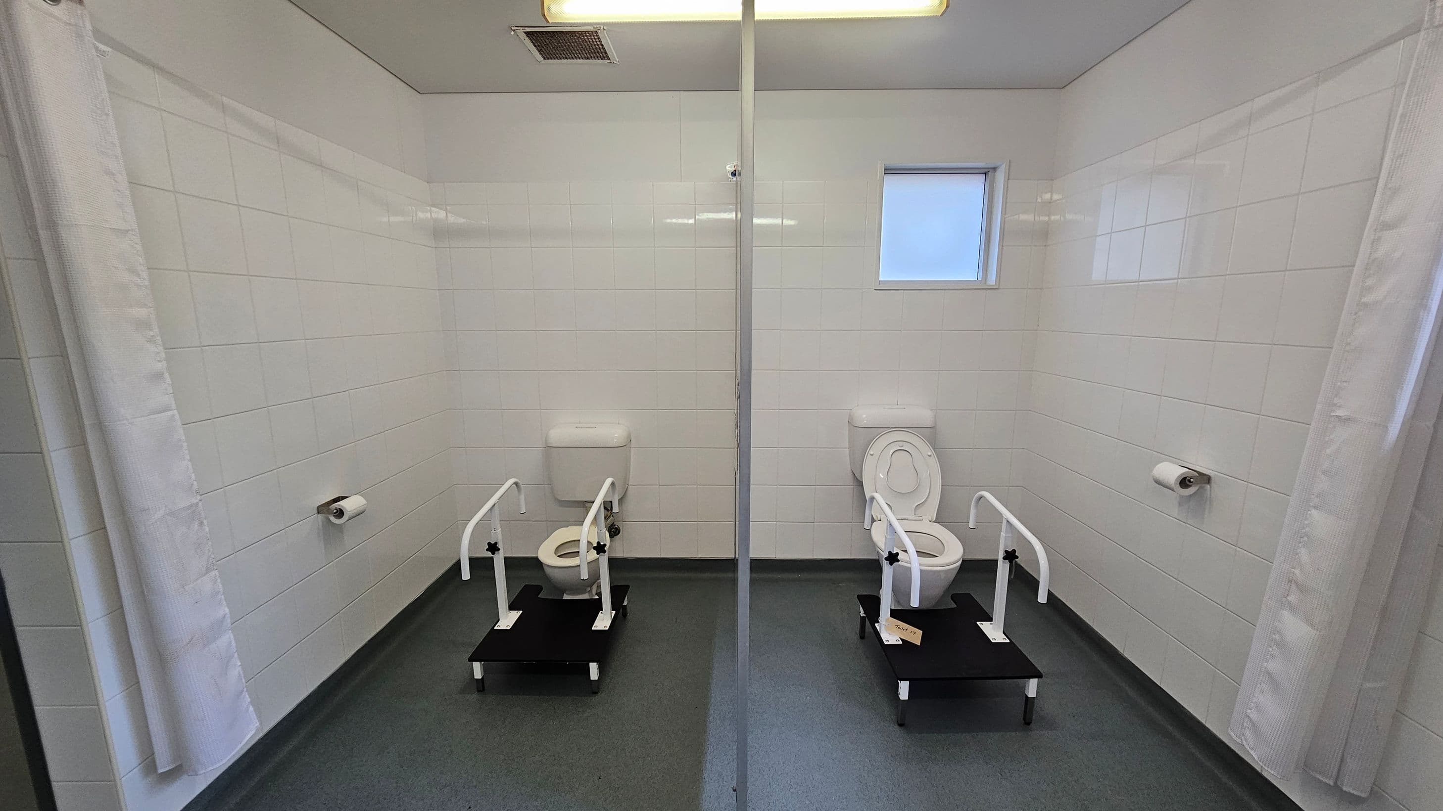 Two toilets are positioned side by side, separated by a glass partition. Each toilet is equipped with support rails and a raised platform. The walls are covered with white tiles, and there is a small window on the right side. Toilet paper holders are mounted on the walls next to each toilet. The floor is grey, and the ceiling is white with a light fixture and vent. A white curtain is partially visible on the left.