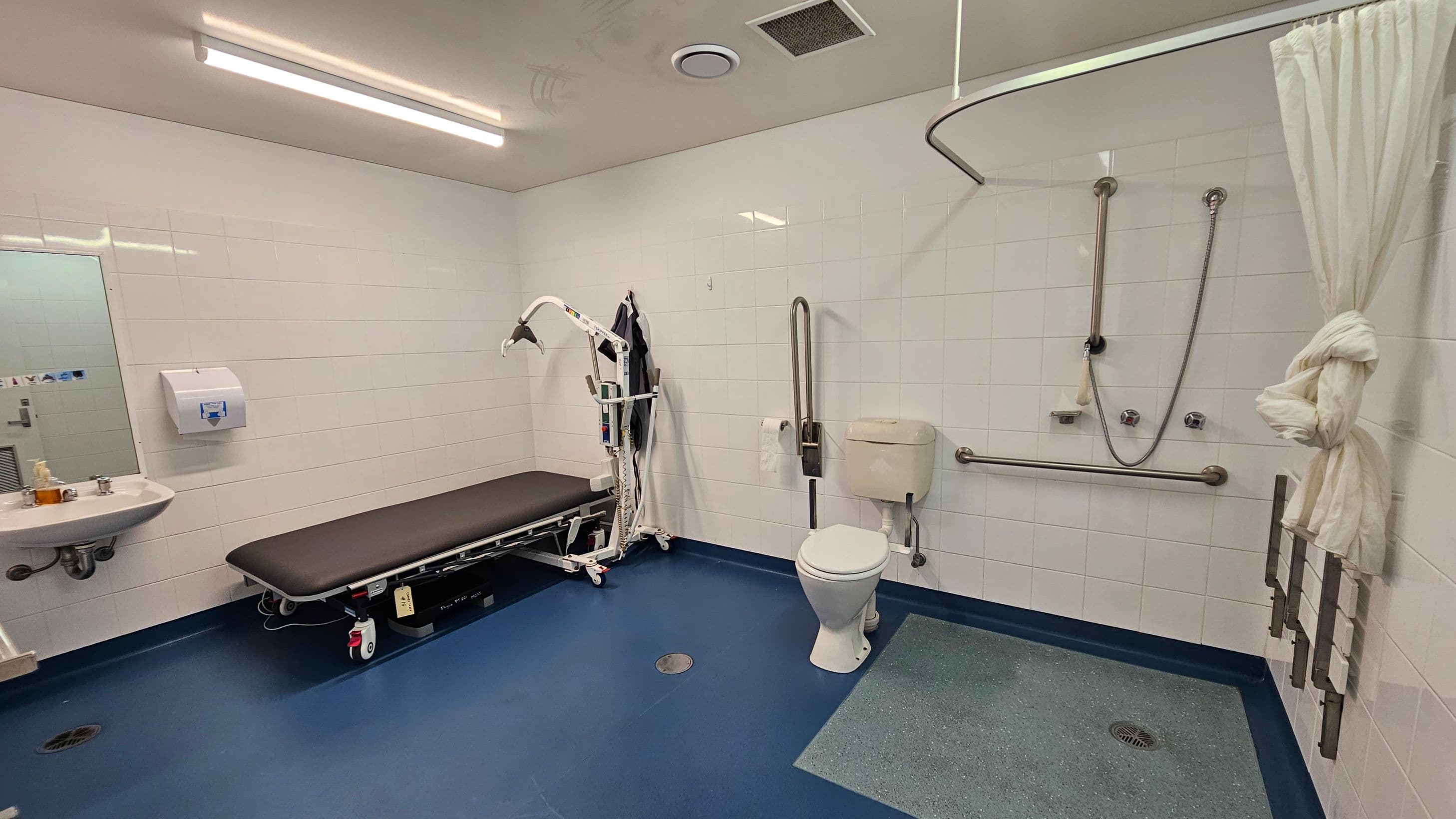 A spacious, accessible bathroom with white tiled walls and a blue floor. On the left, there's a sink with a mirror above it and a paper towel dispenser. A black cushioned bench with a hoist is positioned nearby. On the right, a toilet with grab bars is adjacent to a shower area with a curtain tied to the side. The ceiling has fluorescent lighting and ventilation fixtures.