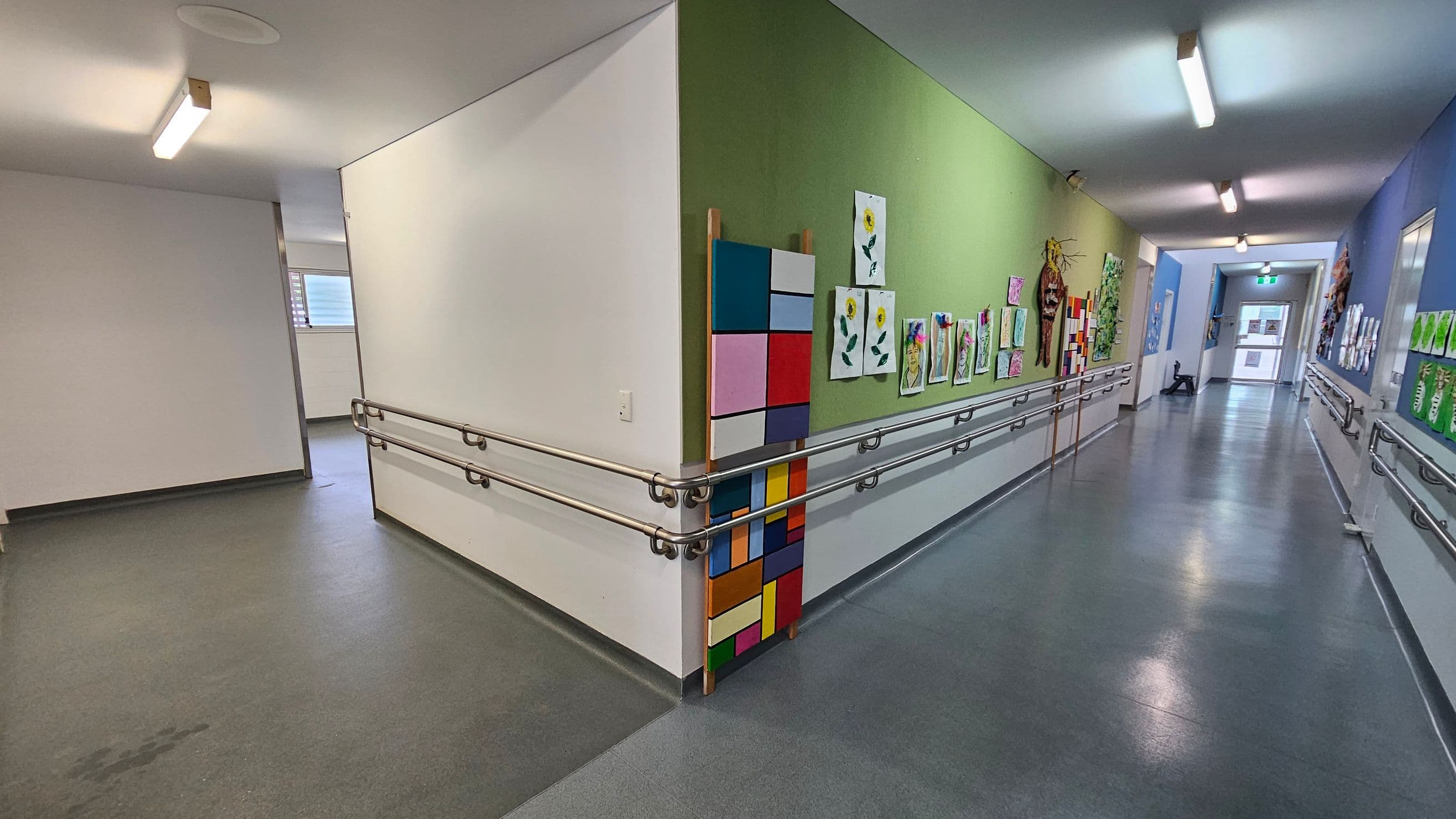 A corridor with a green and blue wall on the right, displaying various colourful artworks, including paintings of flowers and abstract designs. The left side has a plain white wall with a window in the background. The floor is grey, and there are metal handrails along the walls. The ceiling features fluorescent lights. At the end of the corridor, there is a door with a glass panel.