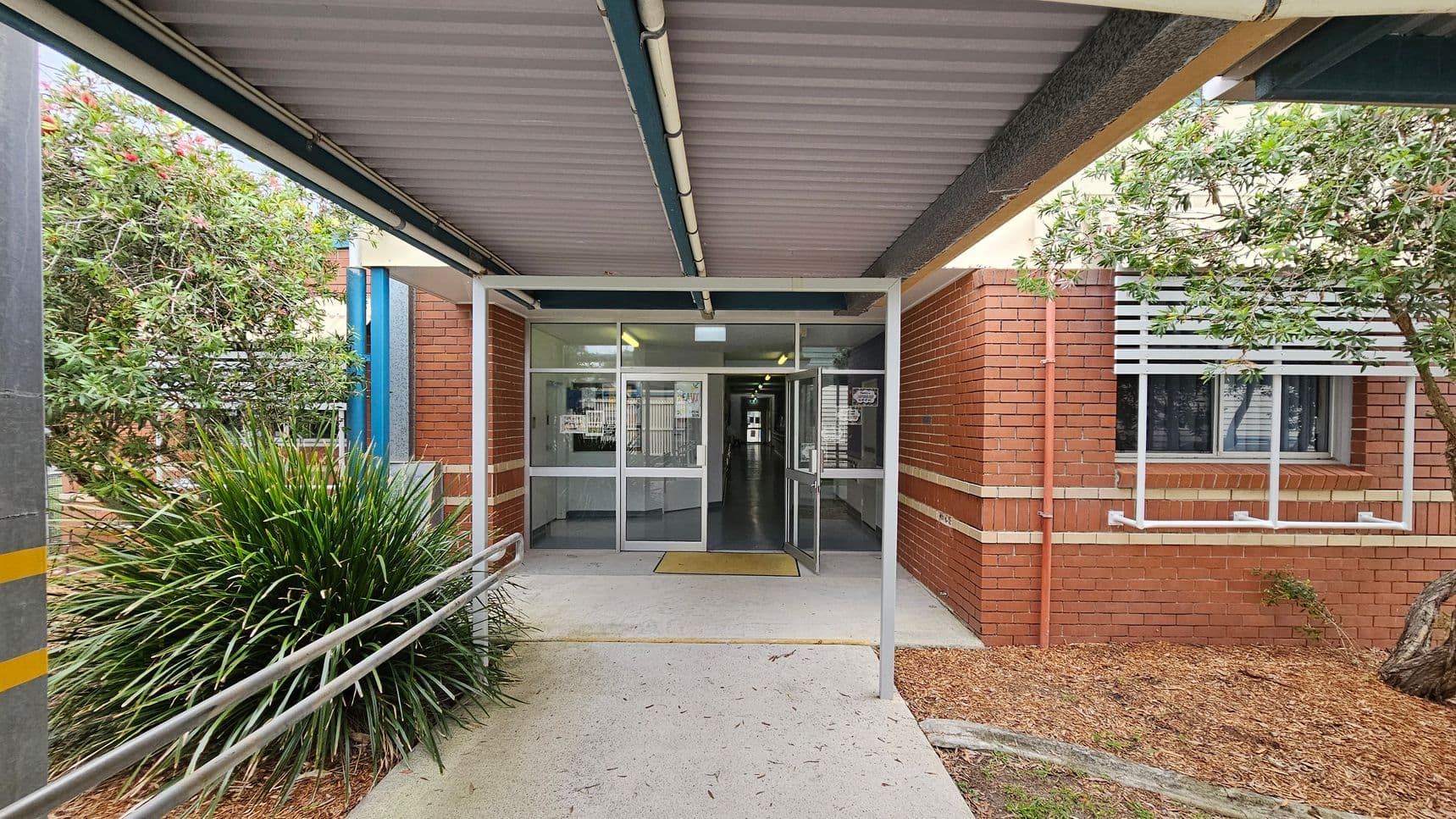 A covered walkway leads to a building entrance with glass doors. The structure has a corrugated metal roof and a metal railing on the left. On the left, there is a bushy plant and a tree, while on the right, a brick wall with a window featuring white slats is visible. The ground is a mix of concrete and mulch. Inside, fluorescent lights illuminate a hallway.