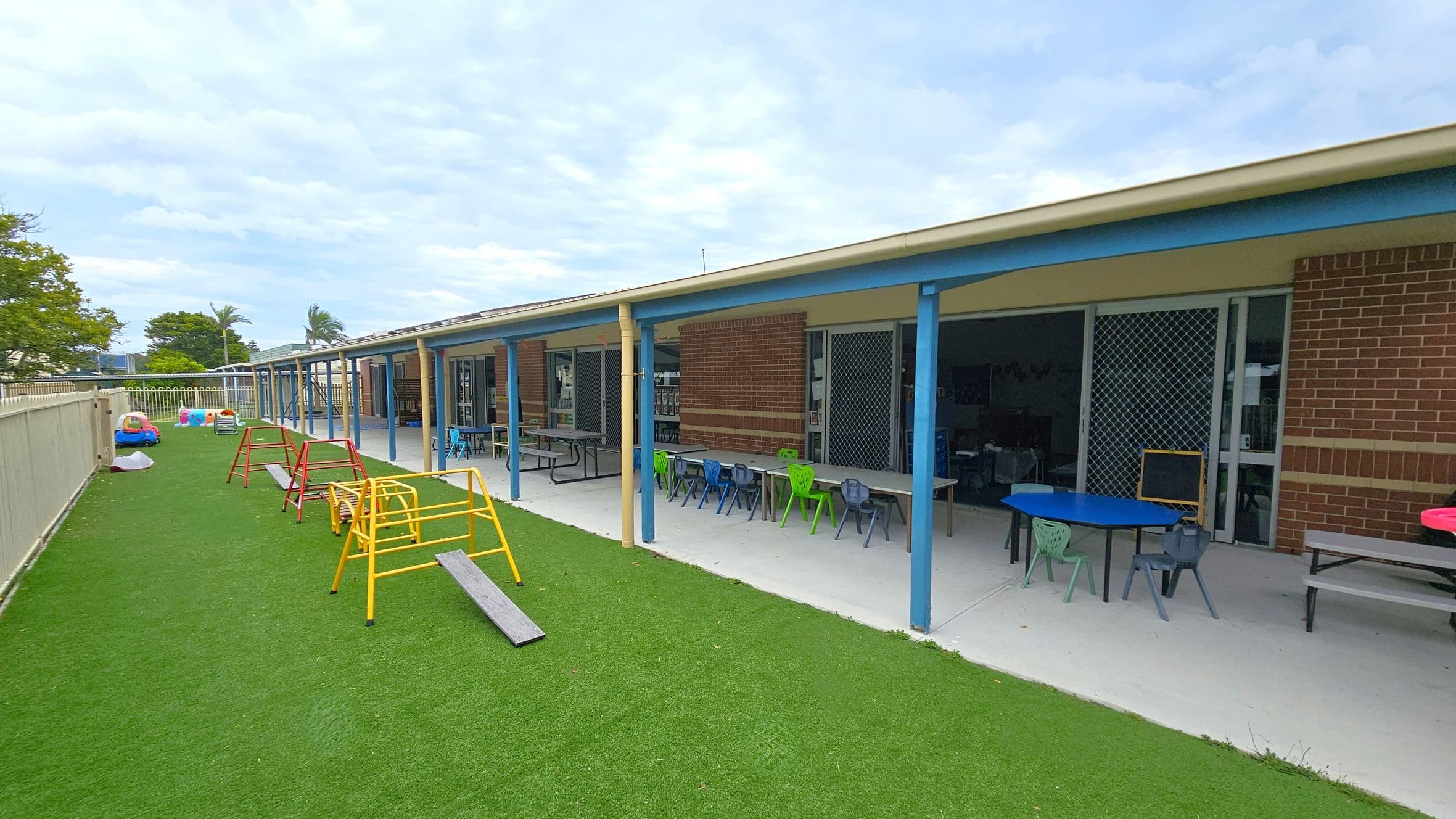 A playground with artificial grass on the left, featuring colourful play equipment like small climbing frames and toy cars. On the right, a building with a covered patio area has tables and chairs in various colours. The building has brick walls and sliding doors with mesh screens. A fence runs along the left side, and there are trees in the background.