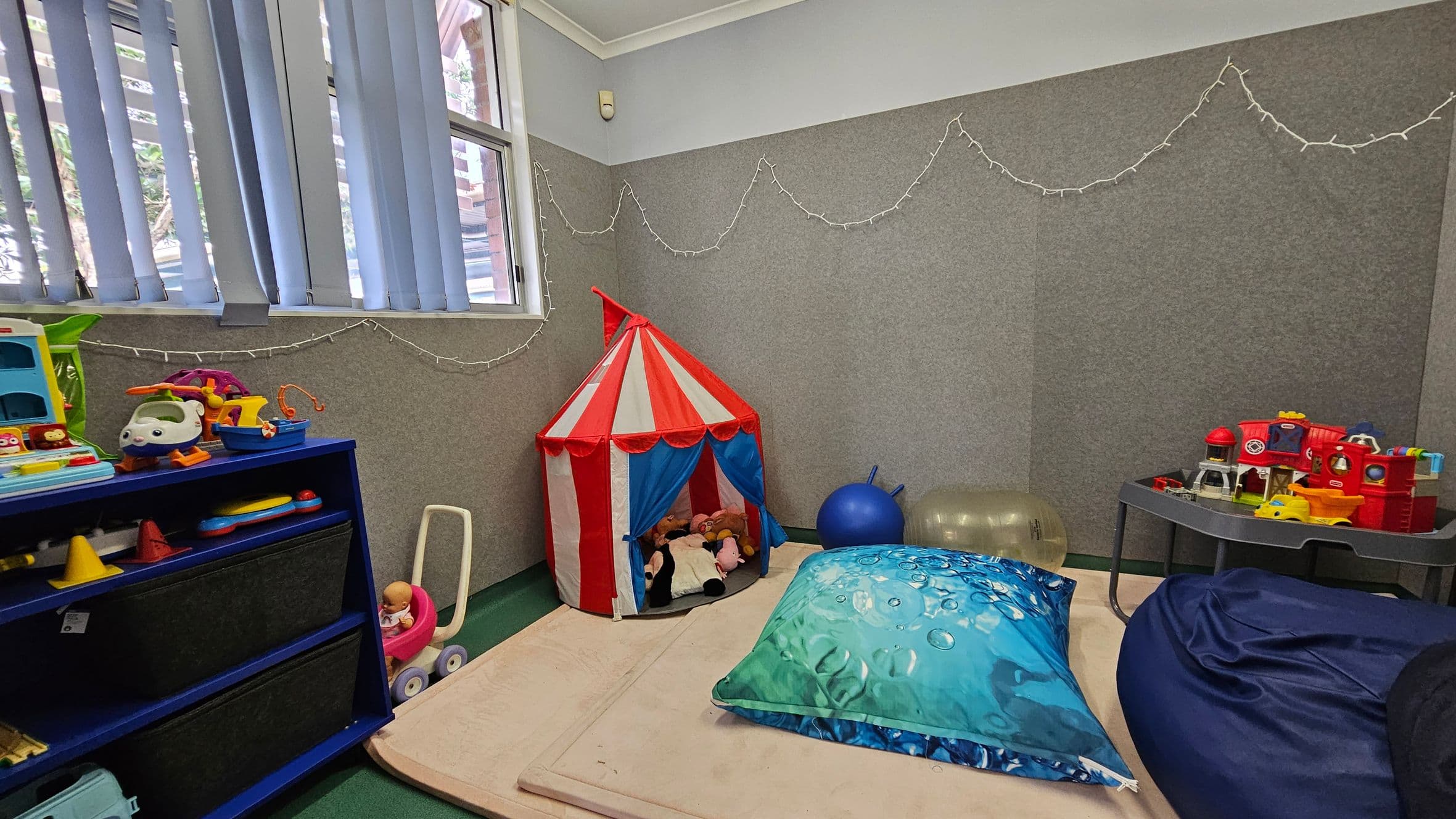 A children's playroom with a green carpeted floor. On the left, a blue shelf holds various toys, including a toy helicopter and a doll in a pink stroller. In the centre, a red and white striped play tent contains stuffed animals. A large blue cushion and a dark blue bean bag are on the floor. On the right, a grey table holds more toys, including a red toy train. The walls are grey with string lights hanging, and there are vertical blinds on the window.