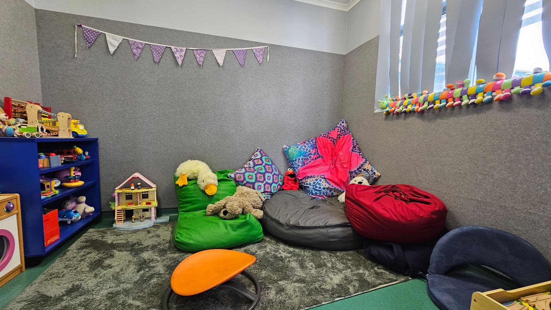 A cosy playroom with grey walls and a green carpet. On the left, a blue shelf holds various toys, including a dollhouse and colourful play items. In the middle, bean bags in green, grey, and red are adorned with plush toys and vibrant cushions. On the right, a window with vertical blinds has a colourful caterpillar toy on the sill. A string of purple bunting hangs on the wall, adding a playful touch. An orange stool is on the floor.