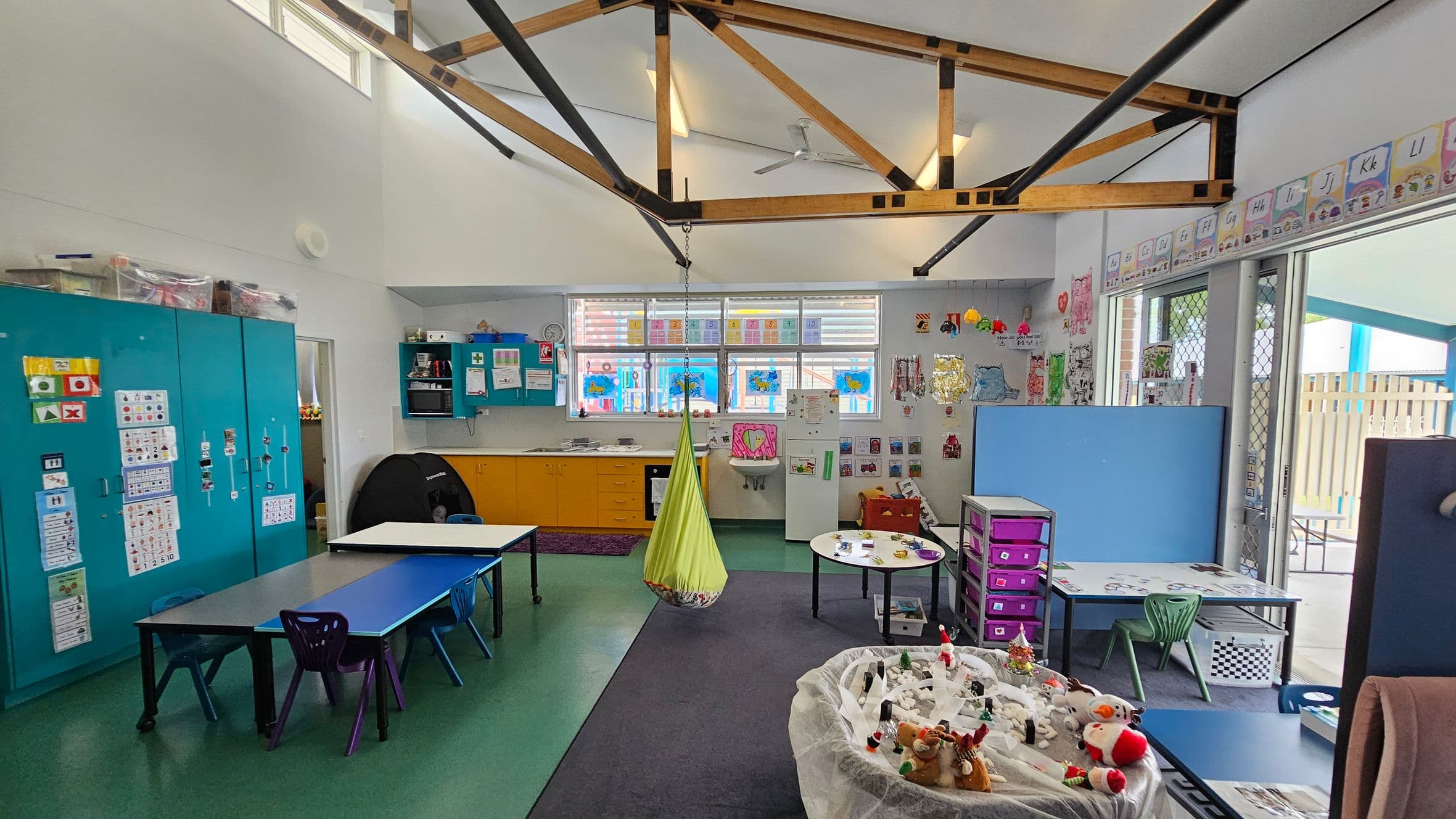 A classroom with a high ceiling featuring exposed wooden beams. On the left, there are blue cabinets with educational posters. In the centre, a green hanging chair is suspended from the ceiling. The back wall has windows with colourful artwork and yellow cabinets below. On the right, tables with chairs and a play area with toys are visible. The floor is a mix of green and grey, and the walls are decorated with various educational materials.