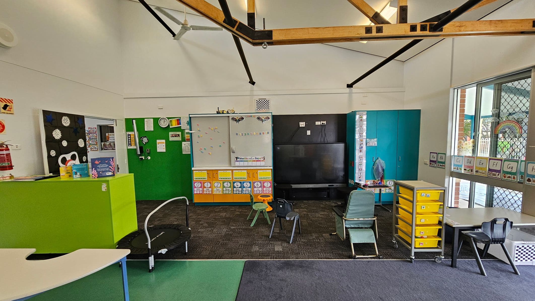 Classroom with a high ceiling featuring exposed wooden beams. The left side has a green partition with educational materials and a fire extinguisher. In the centre, a whiteboard with colourful magnets and a large black TV. Chairs and a small trampoline are on the carpeted floor. The right side has a blue cabinet, a table with yellow drawers, and a window with alphabet cards. The floor is a mix of green and dark carpet.