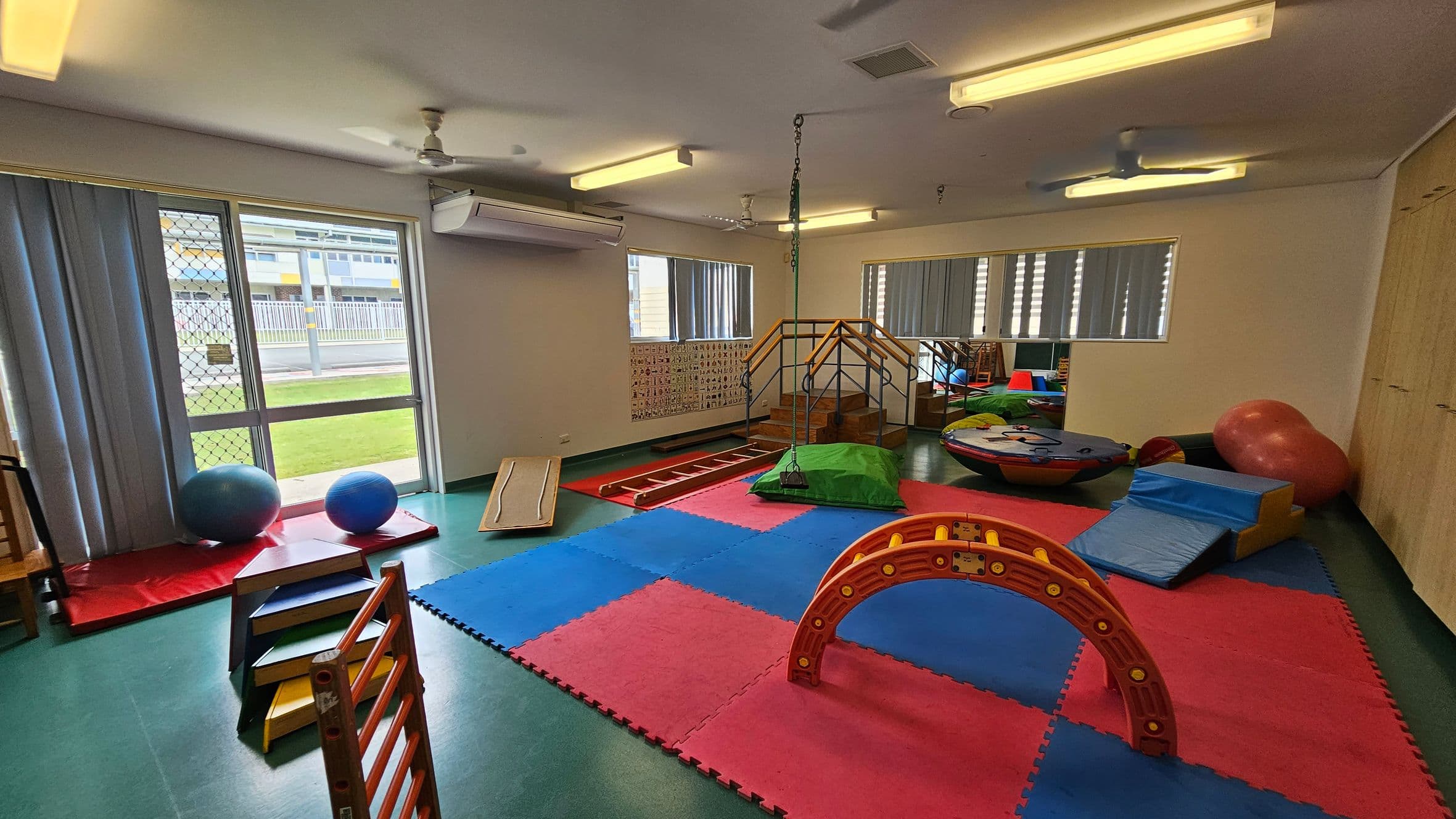A children's playroom with a green floor and a white ceiling. On the left, there are large windows with vertical blinds, a mesh door, and two blue exercise balls on red mats. In the centre, colourful foam mats cover the floor, with a small wooden climbing structure and a red arch. On the right, there's a large red exercise ball, blue foam blocks, and a circular play platform. The back wall has a small staircase with a railing and a chain hanging from the ceiling.